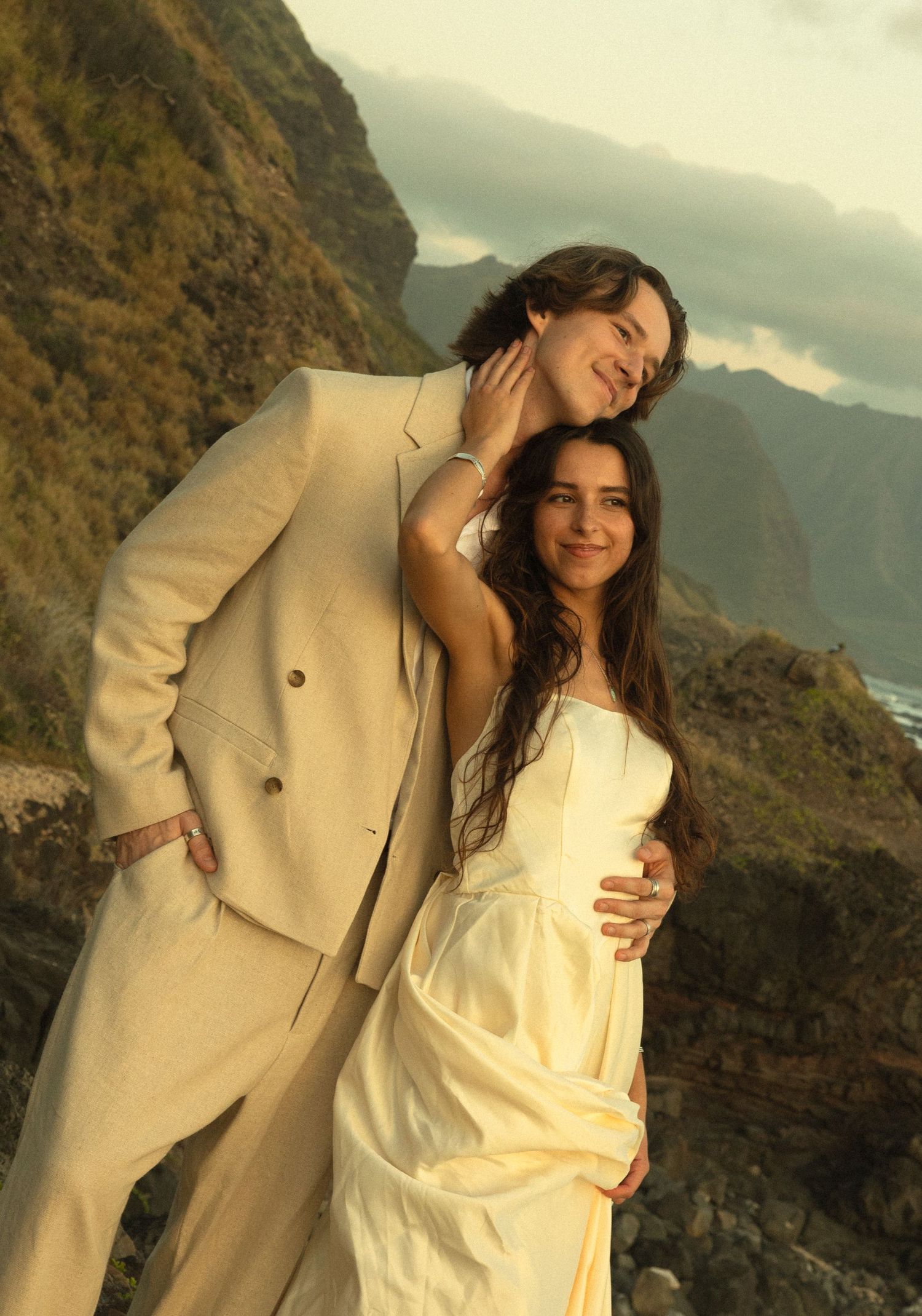 A couple in light-colored formal wear embrace on a scenic mountain cliff overlooking the ocean at sunset.