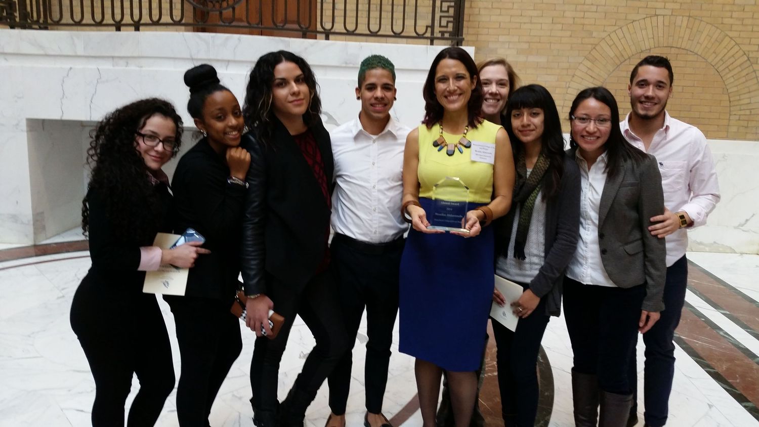 A group of professionals in business attire standing together and smiling in front of a beige brick wall.