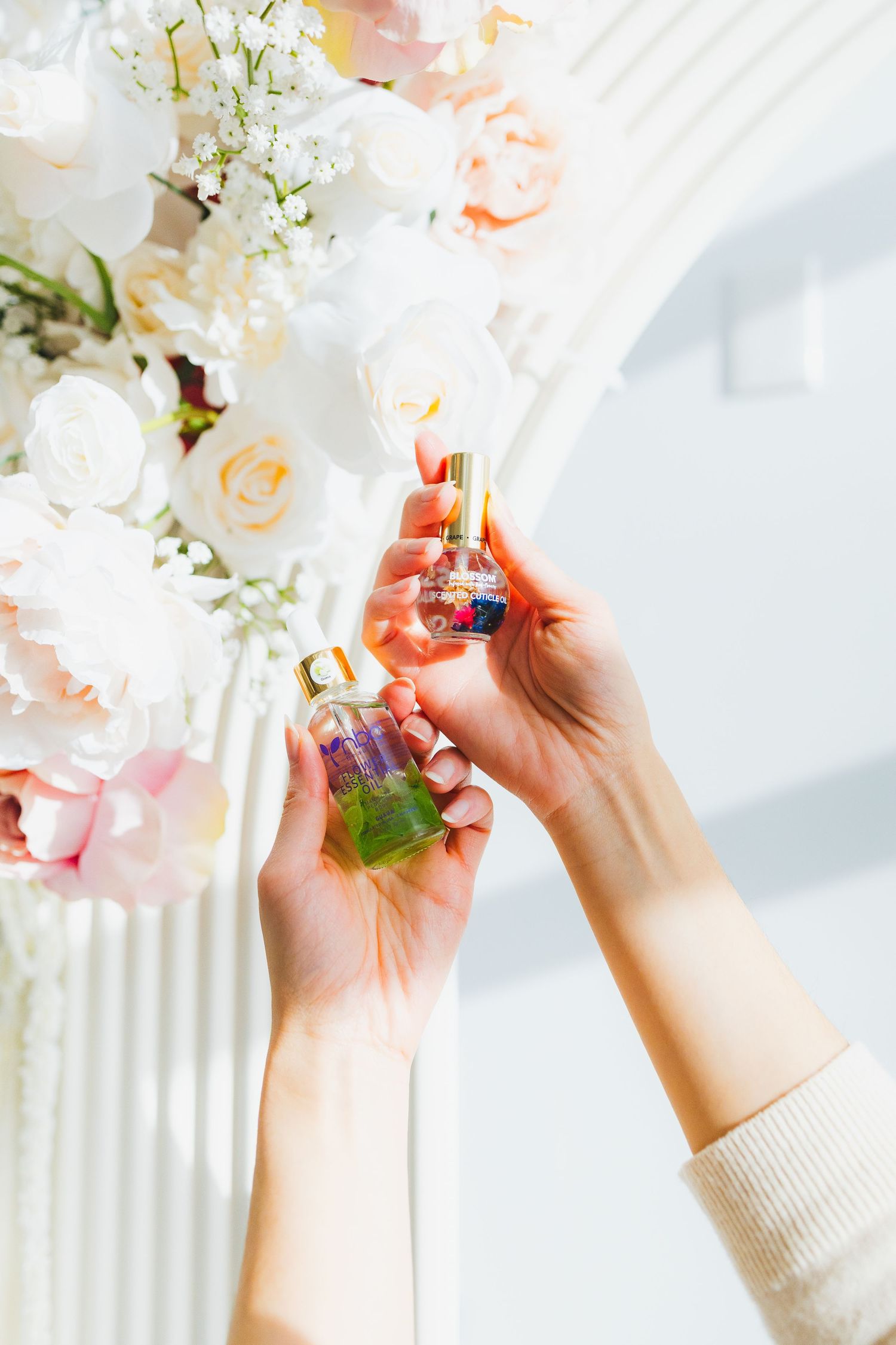 Hands holding perfume bottle against backdrop of soft pink and white floral arrangements.