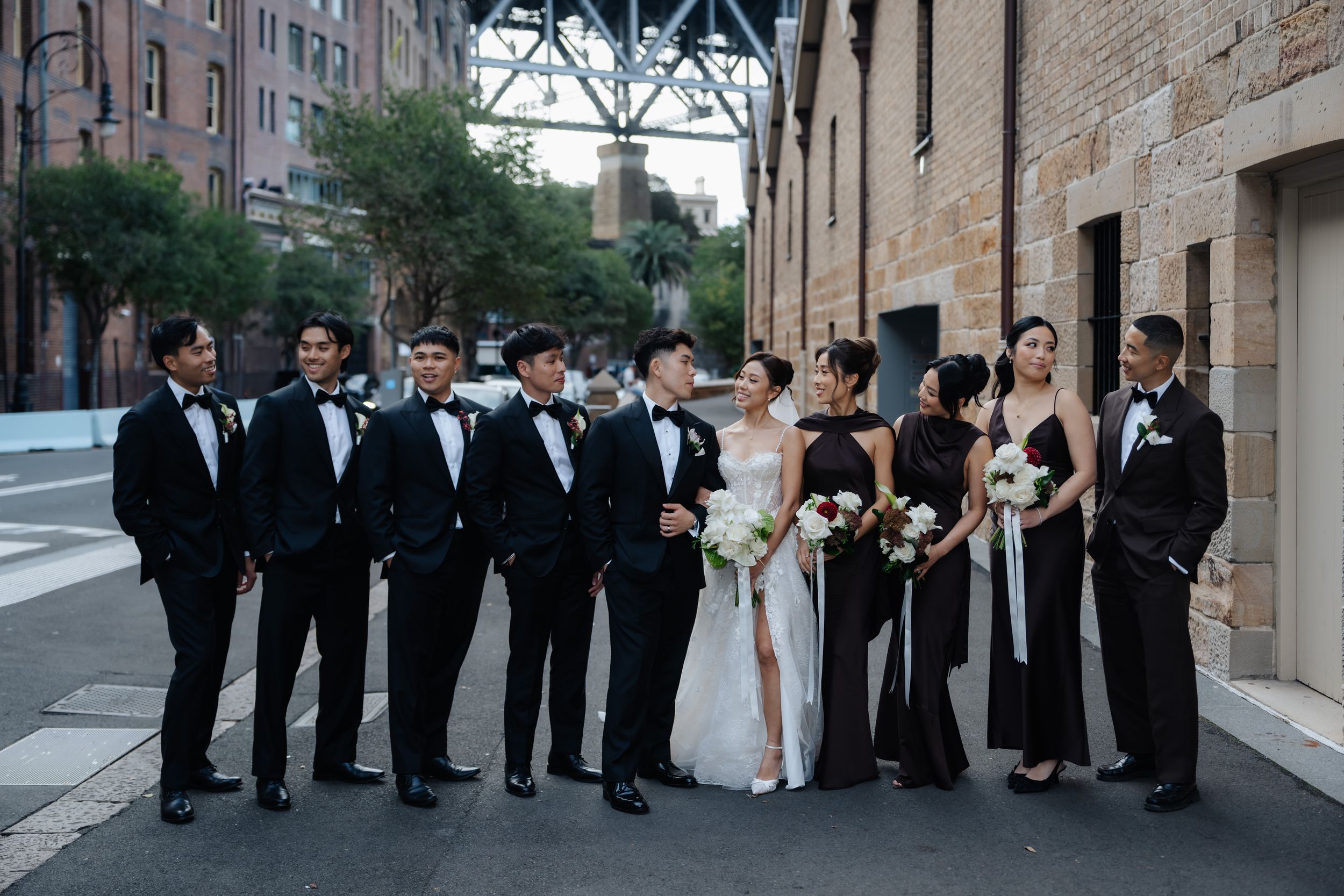 Wedding party in black tuxedos and gowns posing on urban street with bride in white gown holding bouquet.