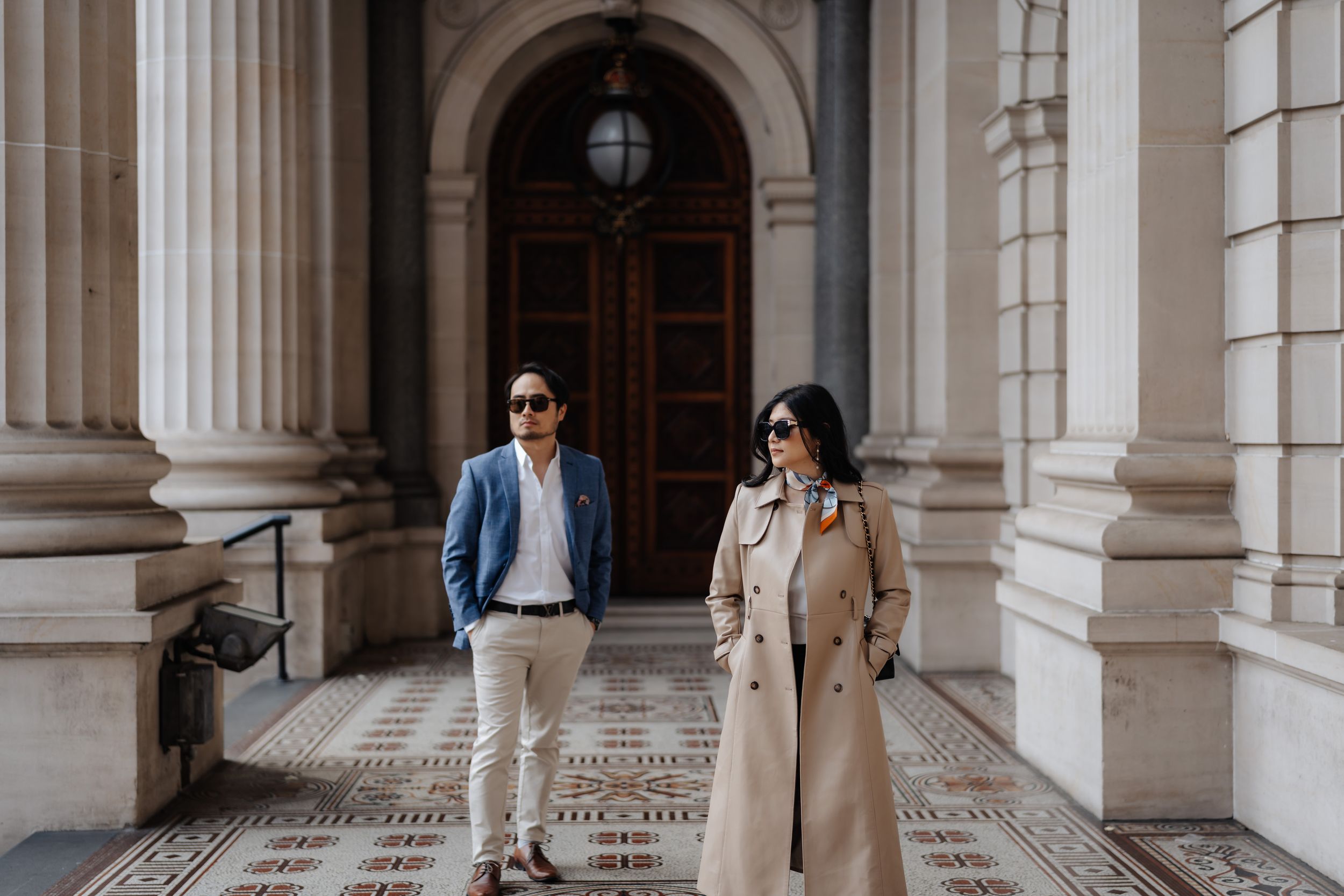 Elegant couple posing outside a grand building with ornate columns, man in blue blazer, woman in beige trench coat.