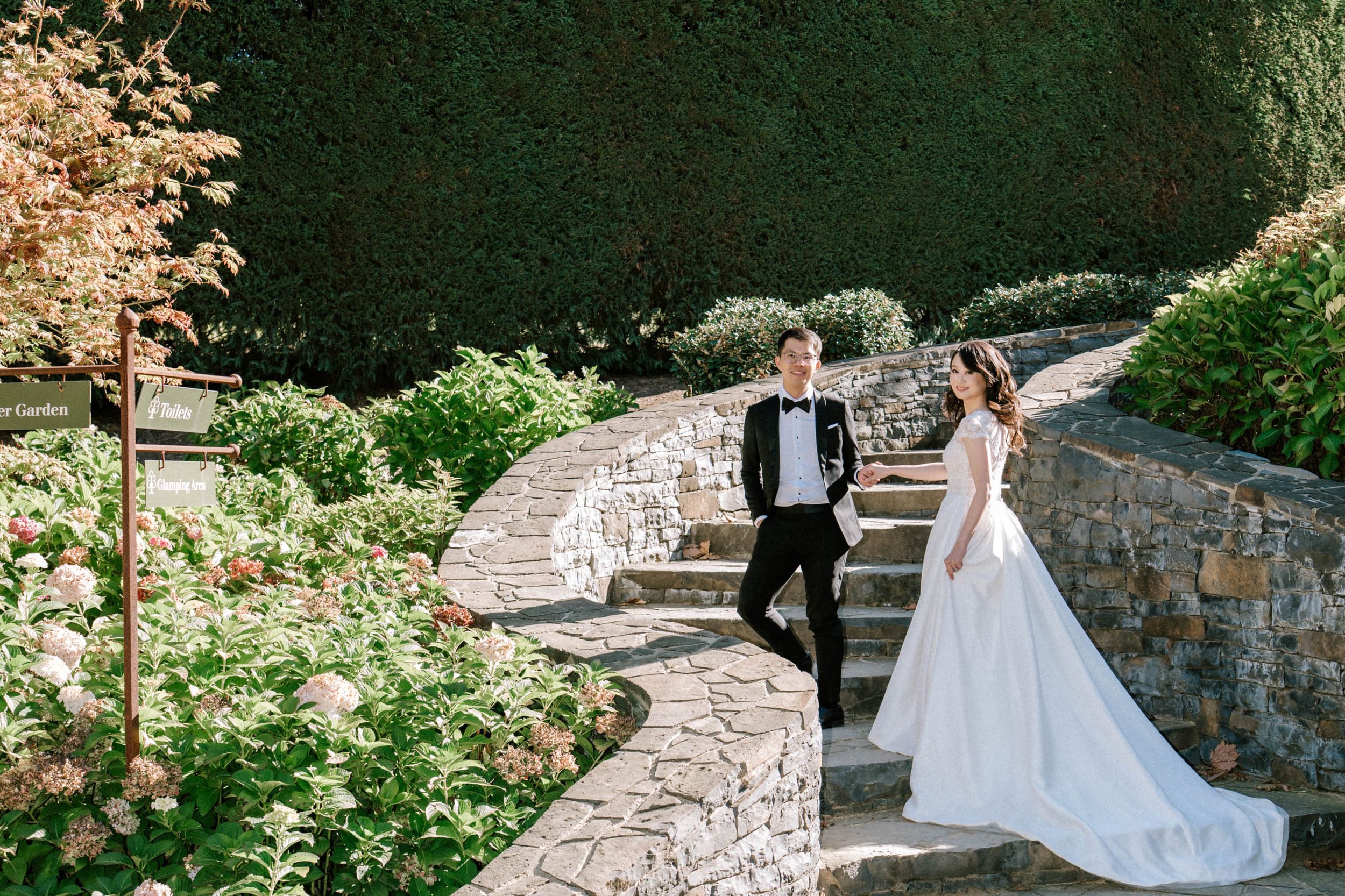 Wedding Photography Sydney - A couple in wedding attire walk together up rustic stone garden steps surrounded by lush greenery and flowers.