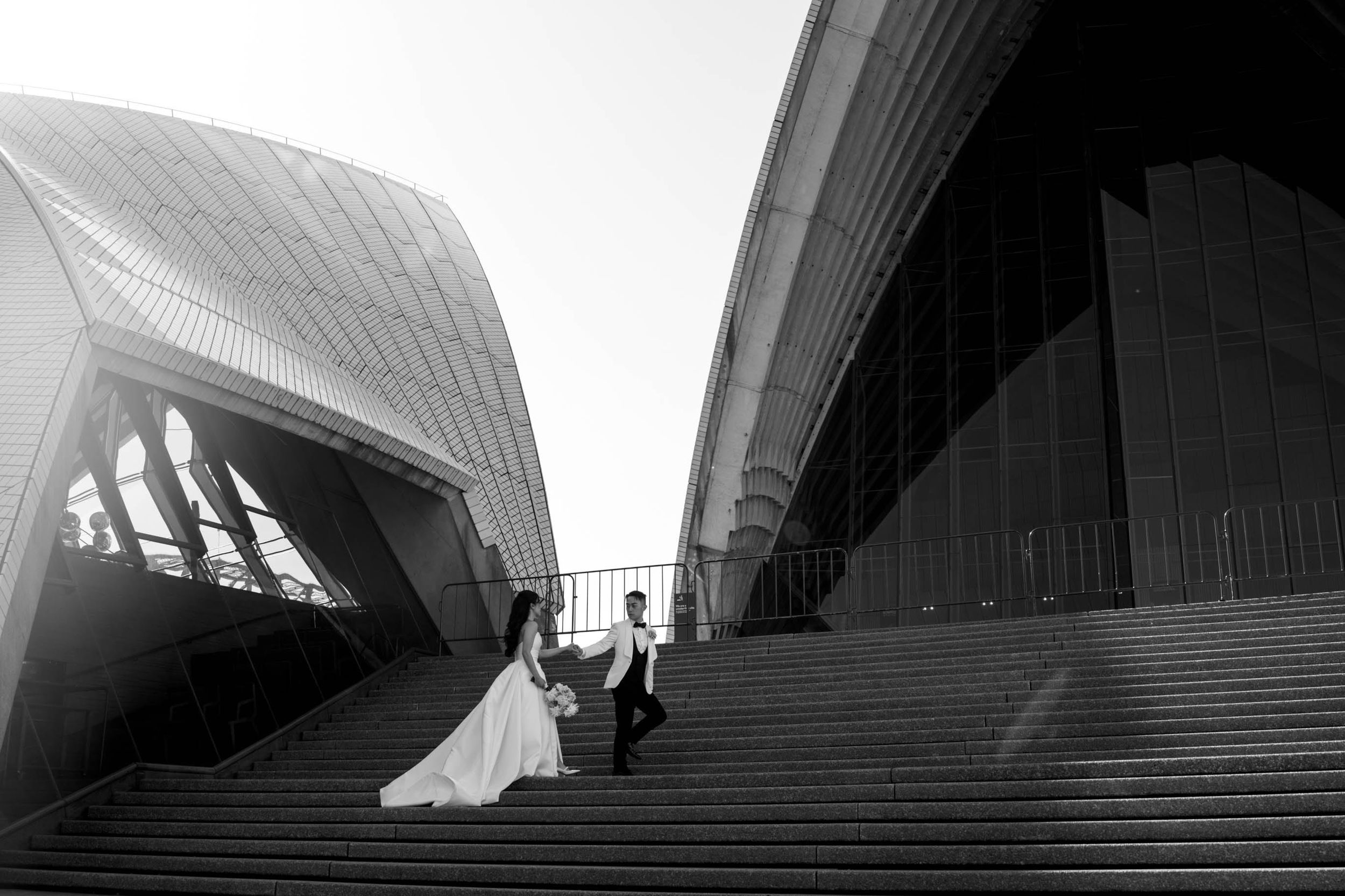 Wedding Photography Sydney - Two Peaches Photography. A couple stands on the grand stone steps of the Sydney Opera House in black and white.