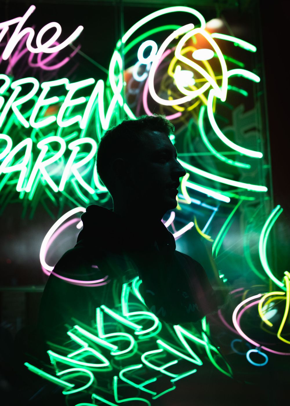 Silhouetted figure stands against vibrant neon signs displaying colorful text and shapes in green and pink hues.
