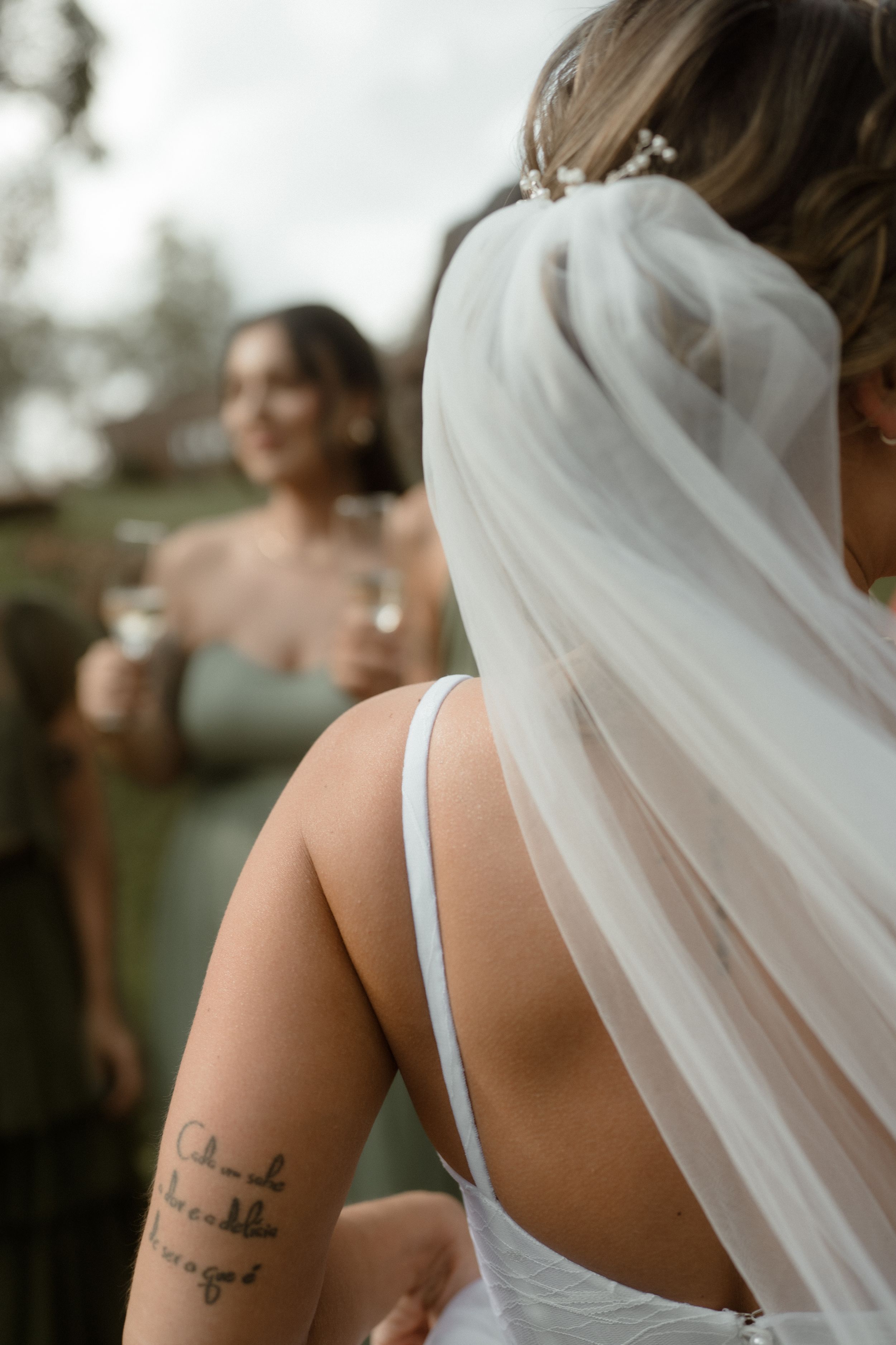 A bride in a white dress with a flowing veil shows her arm tattoo while someone in sage green stands in the background.
