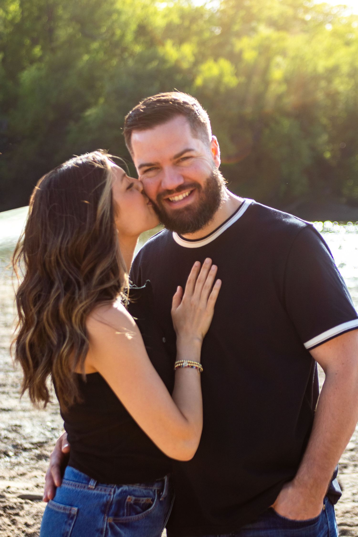 A woman kisses a smiling bearded man on the cheek outdoors in golden hour light.