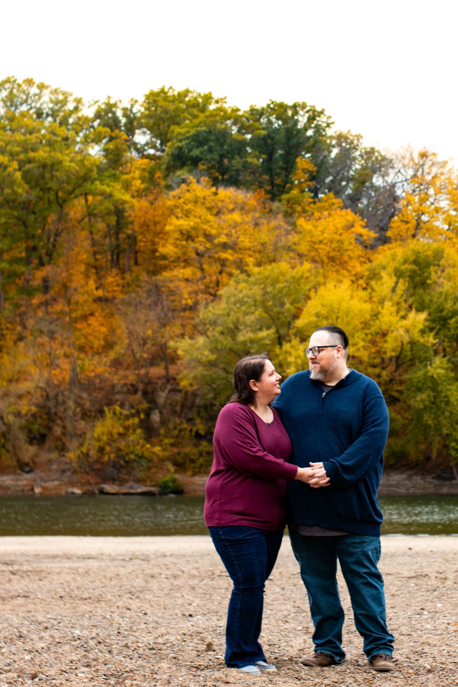 Couple holding hands and gazing at each other on a rocky shore with vibrant autumn foliage and a river backdrop.