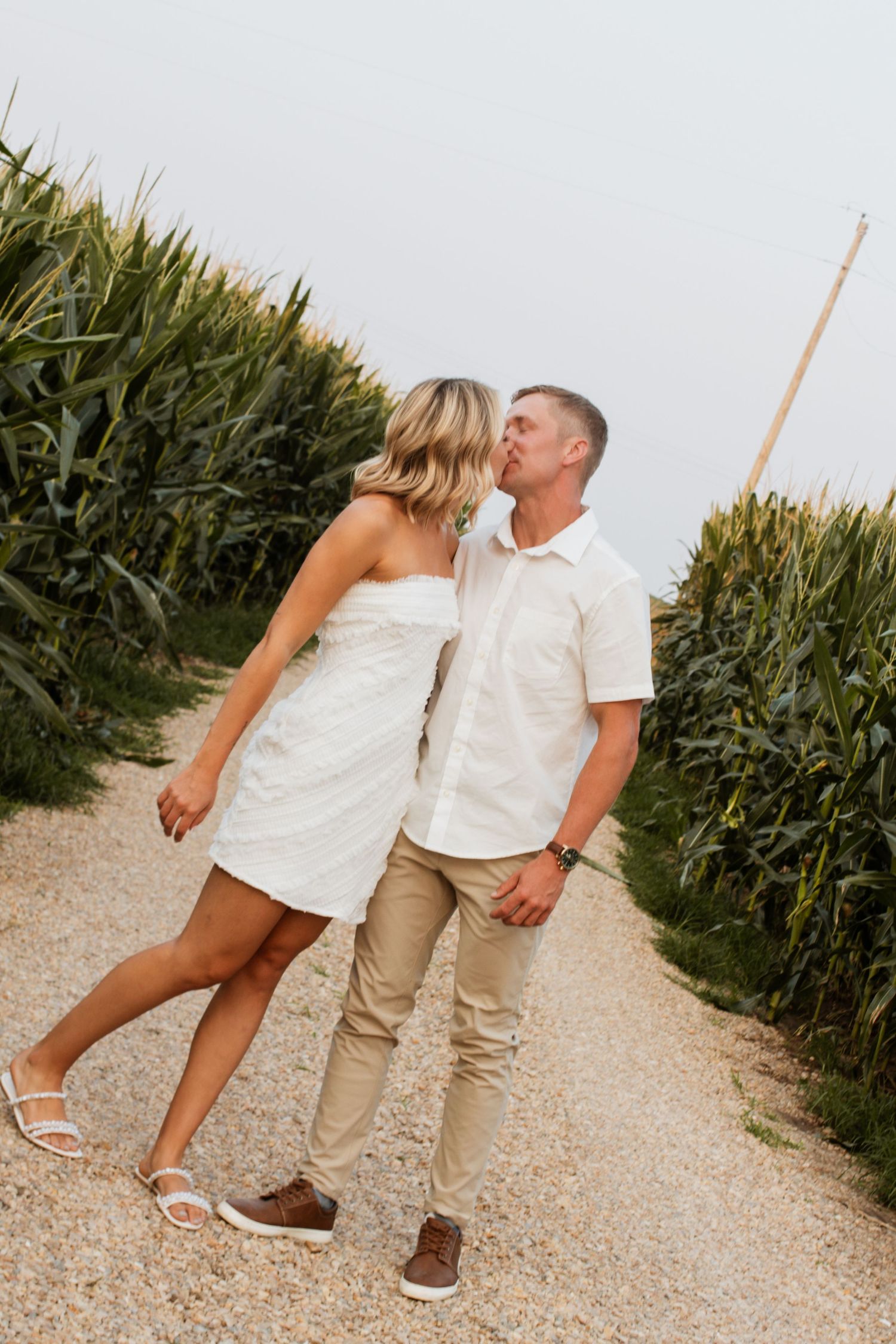Romantic couple in white outfits walking through a corn maze pathway on a sunny day.