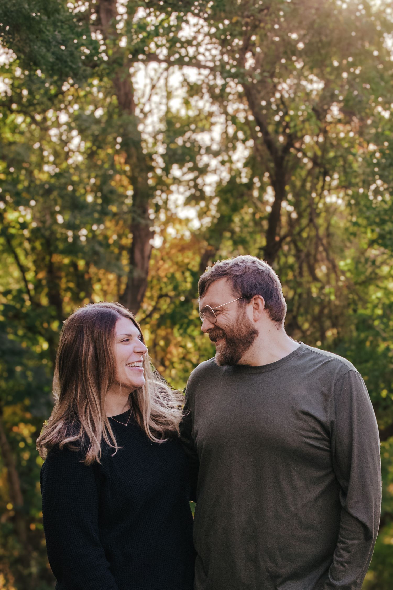 A smiling couple stands together in a sunlit forest, the woman in black and man in olive green sharing a warm glance.