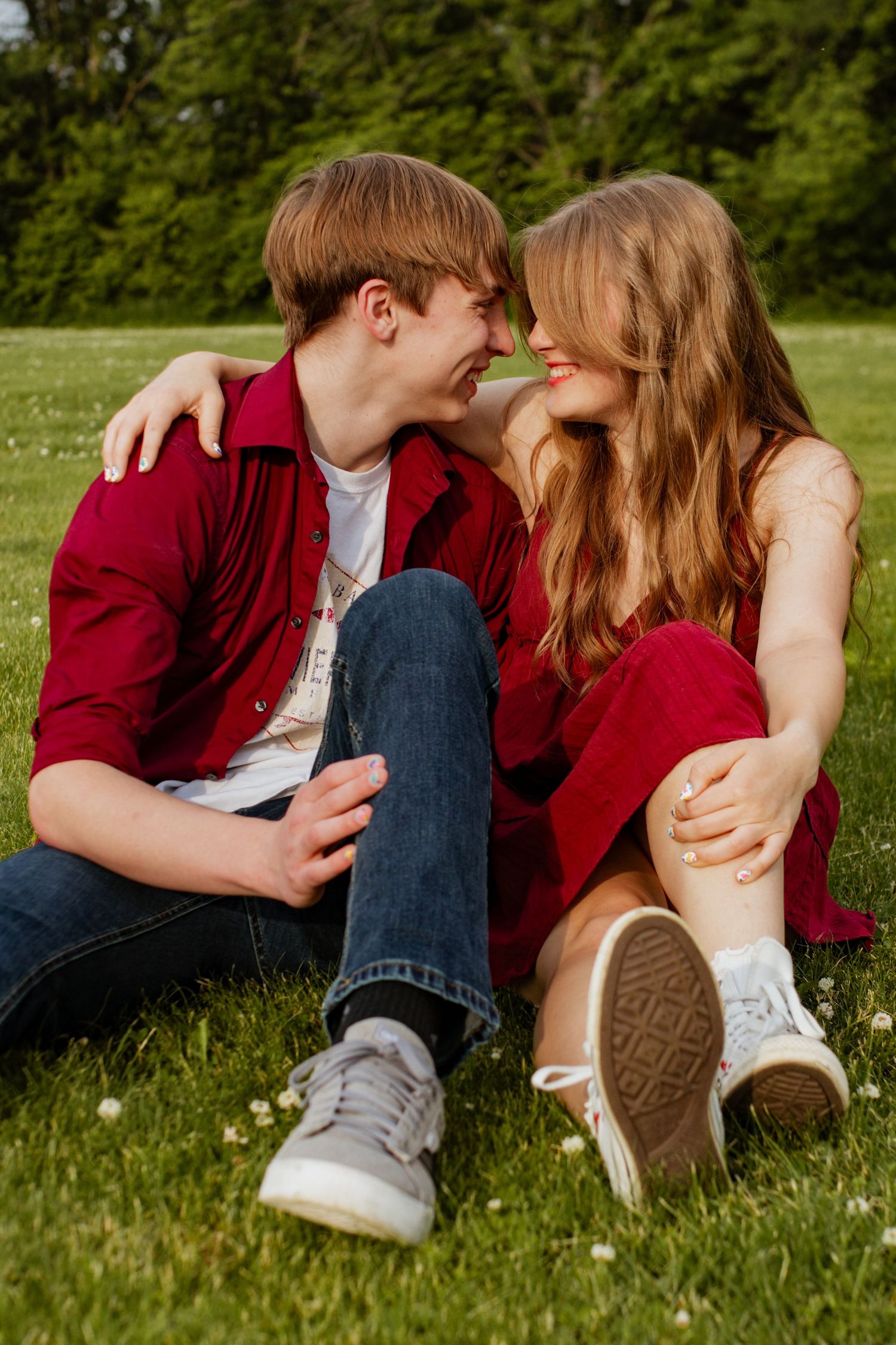 A young couple in matching red outfits sits on green grass, smiling and leaning in for a kiss outdoors.