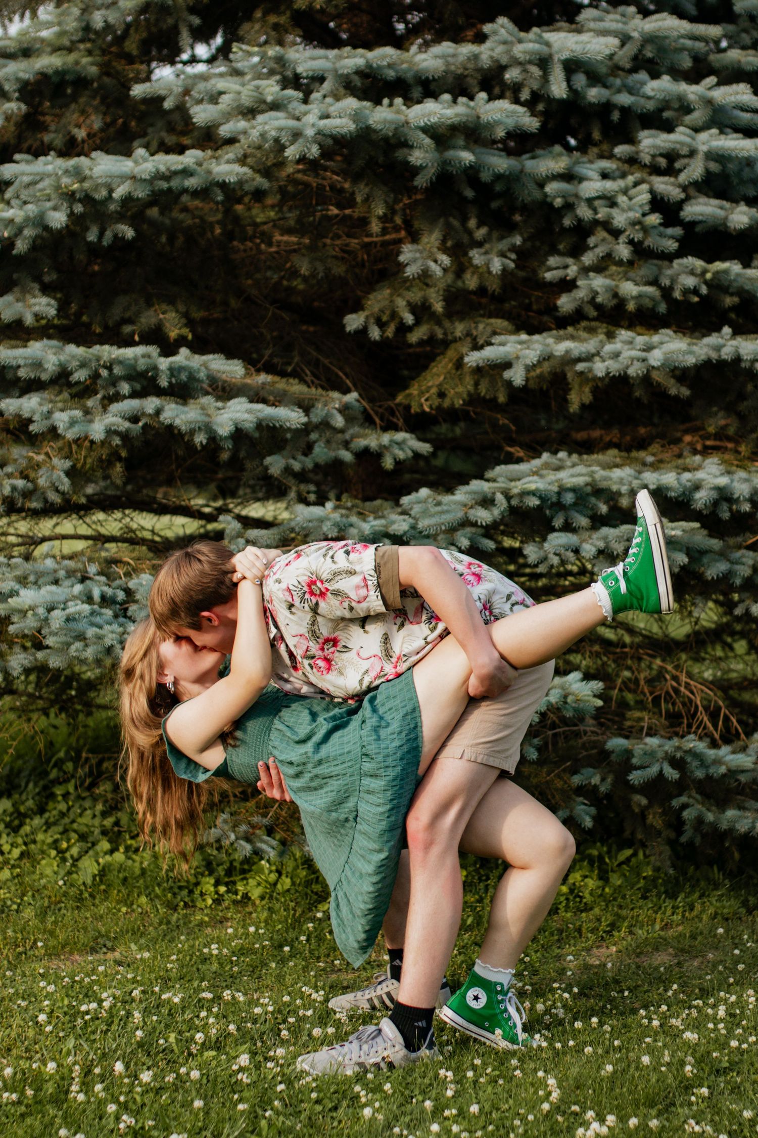Couple sharing a romantic dip kiss in a green park with blue spruce trees, woman wearing floral top and green Converse.