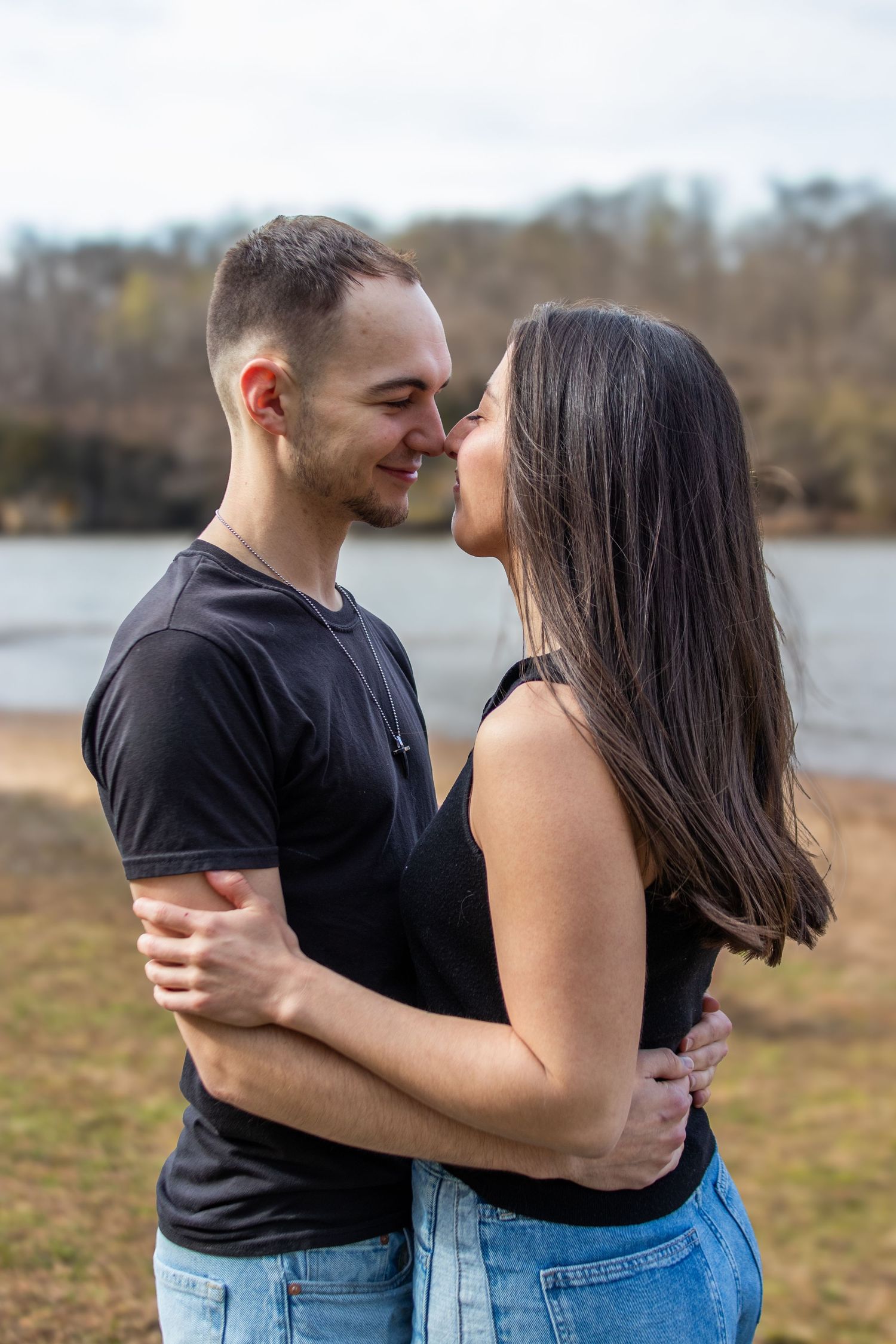 A young couple in matching black tops and jeans share a romantic moment near a lake, noses touching and smiling.