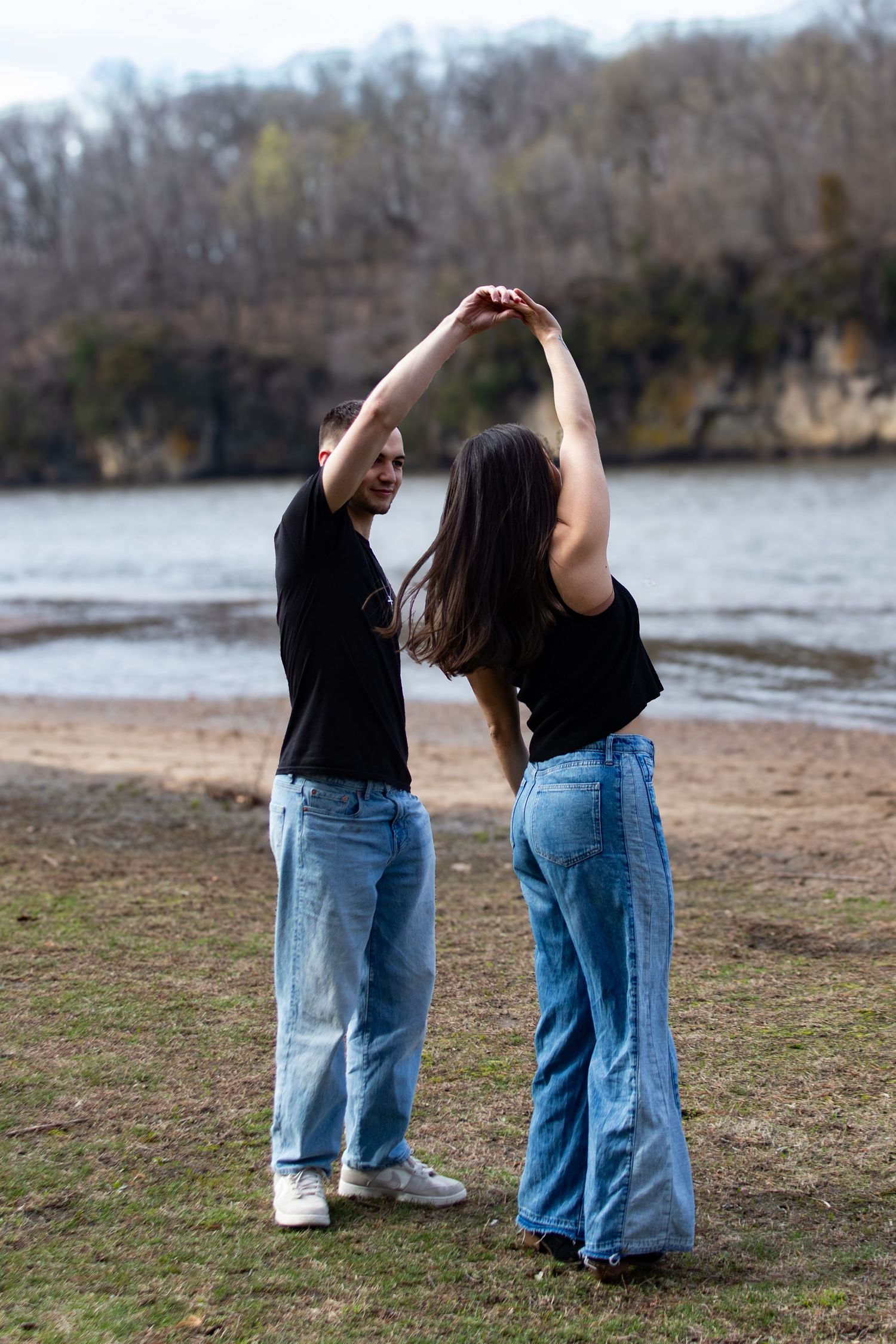 Couple dancing outdoors near a river, both wearing black tops and wide-leg jeans, holding hands raised above.