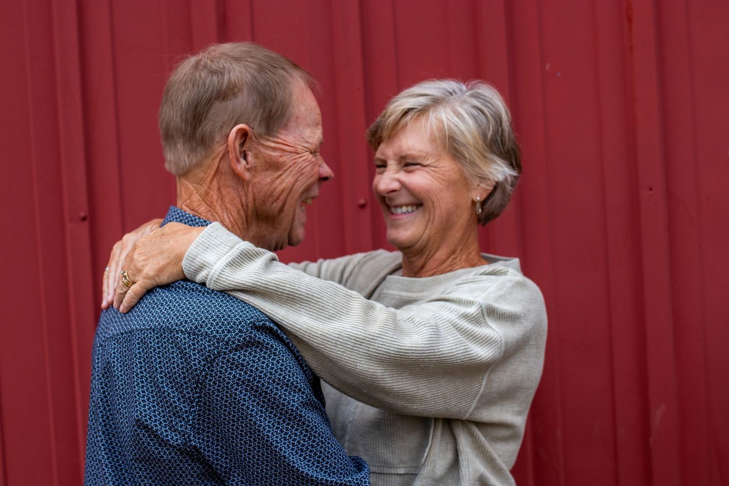 Elderly couple smiling and embracing each other affectionately in front of a red barn wall.