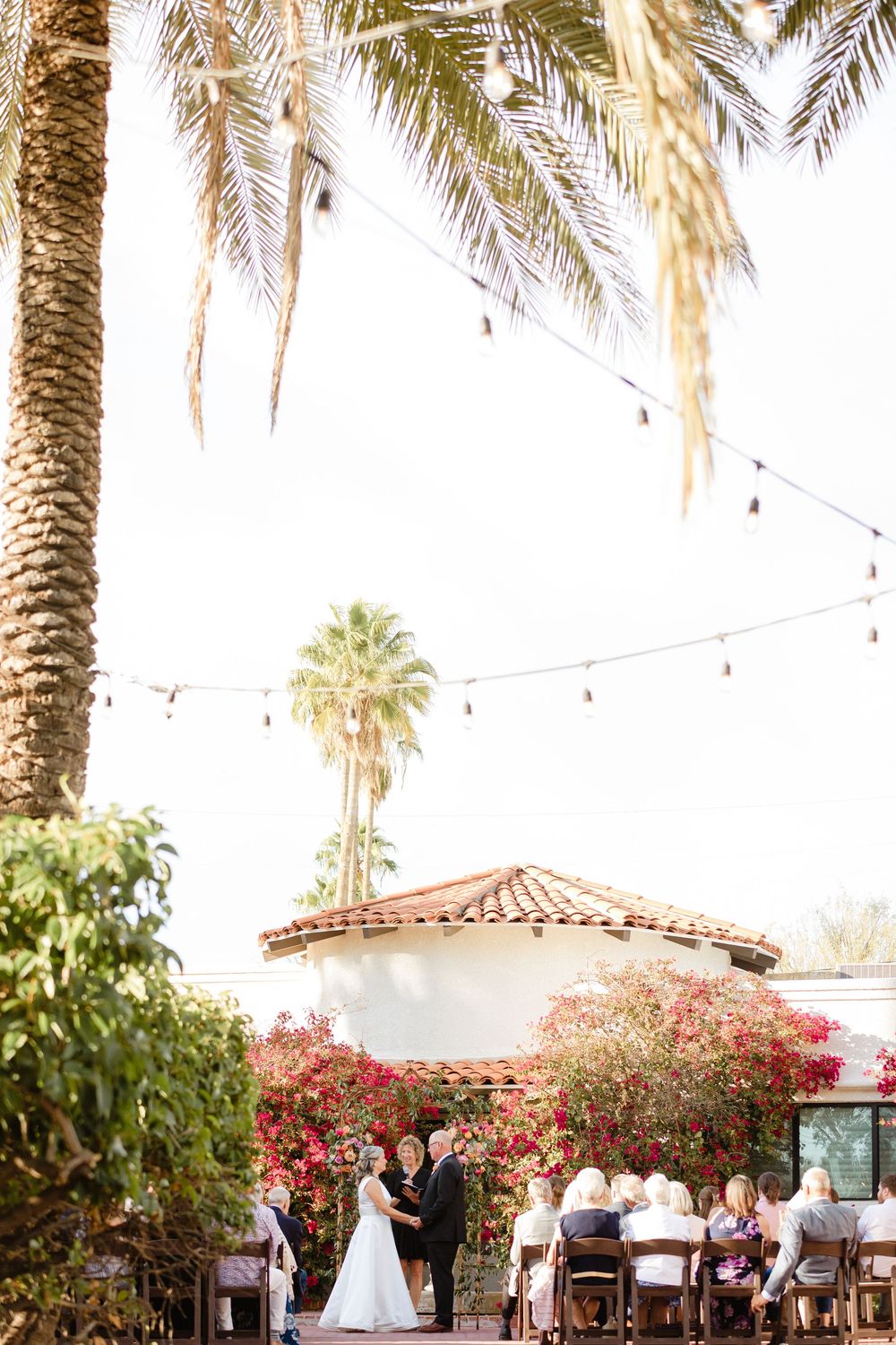 Outdoor wedding ceremony at a Spanish-style venue with palm trees, string lights, and blooming bougainvillea flowers.