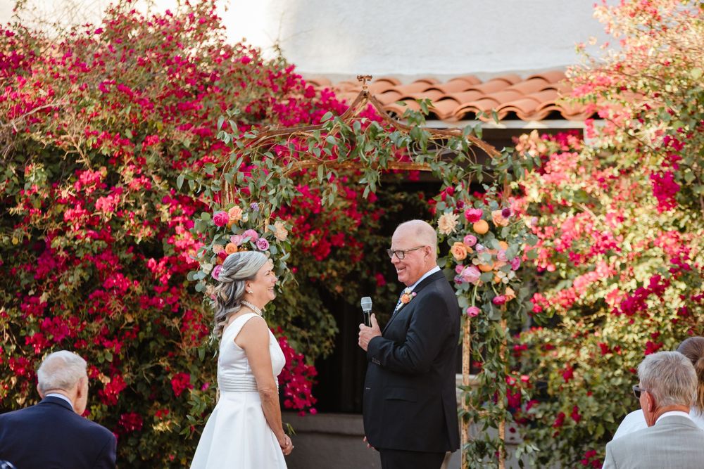 An outdoor wedding ceremony takes place under a vibrant archway of blooming red bougainvillea flowers.