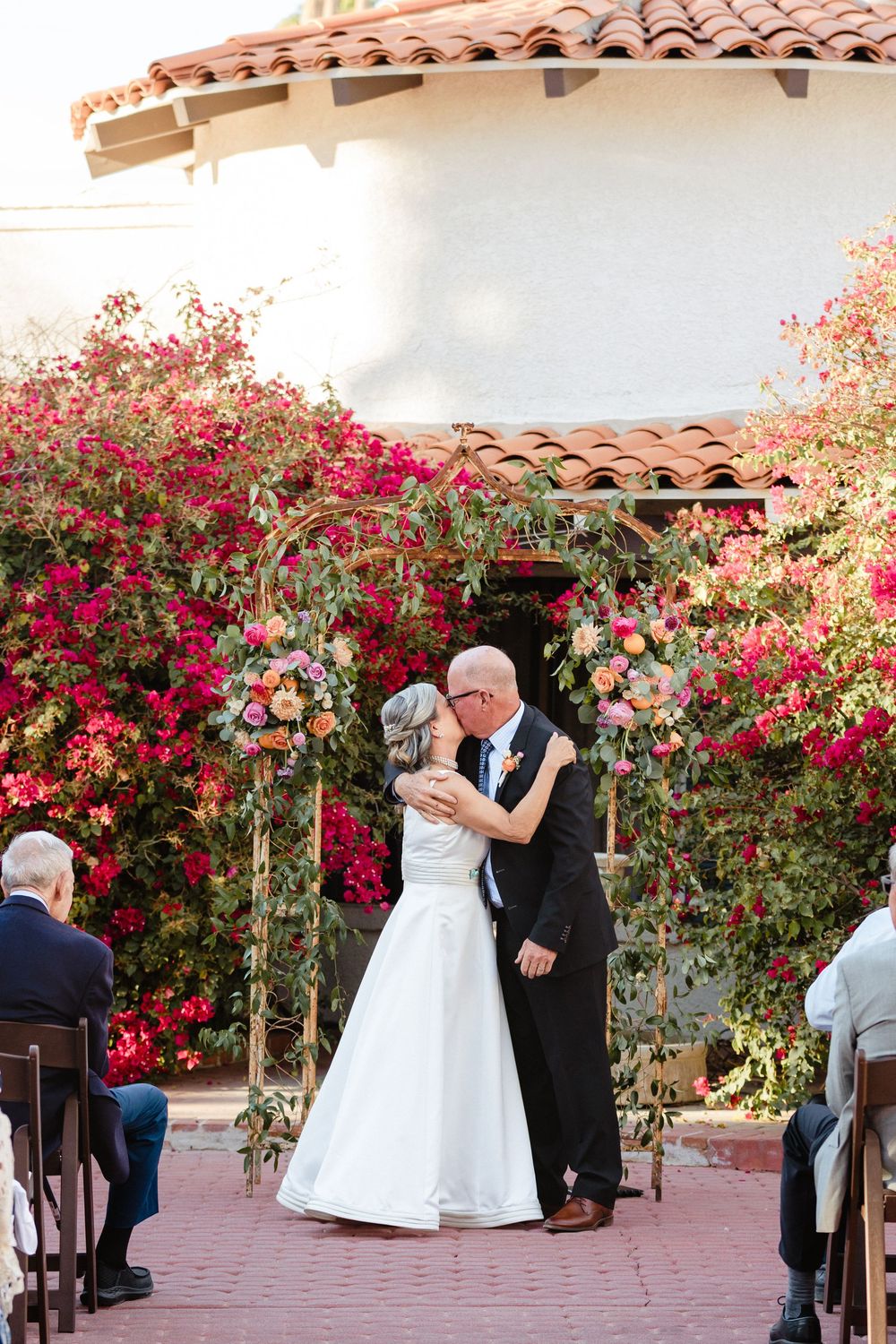 An intimate outdoor wedding moment beneath a flower-covered archway with terra cotta roof tiles overhead.