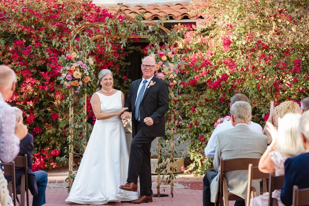 An outdoor wedding ceremony takes place under a flowering red bougainvillea arbor in a garden courtyard setting.