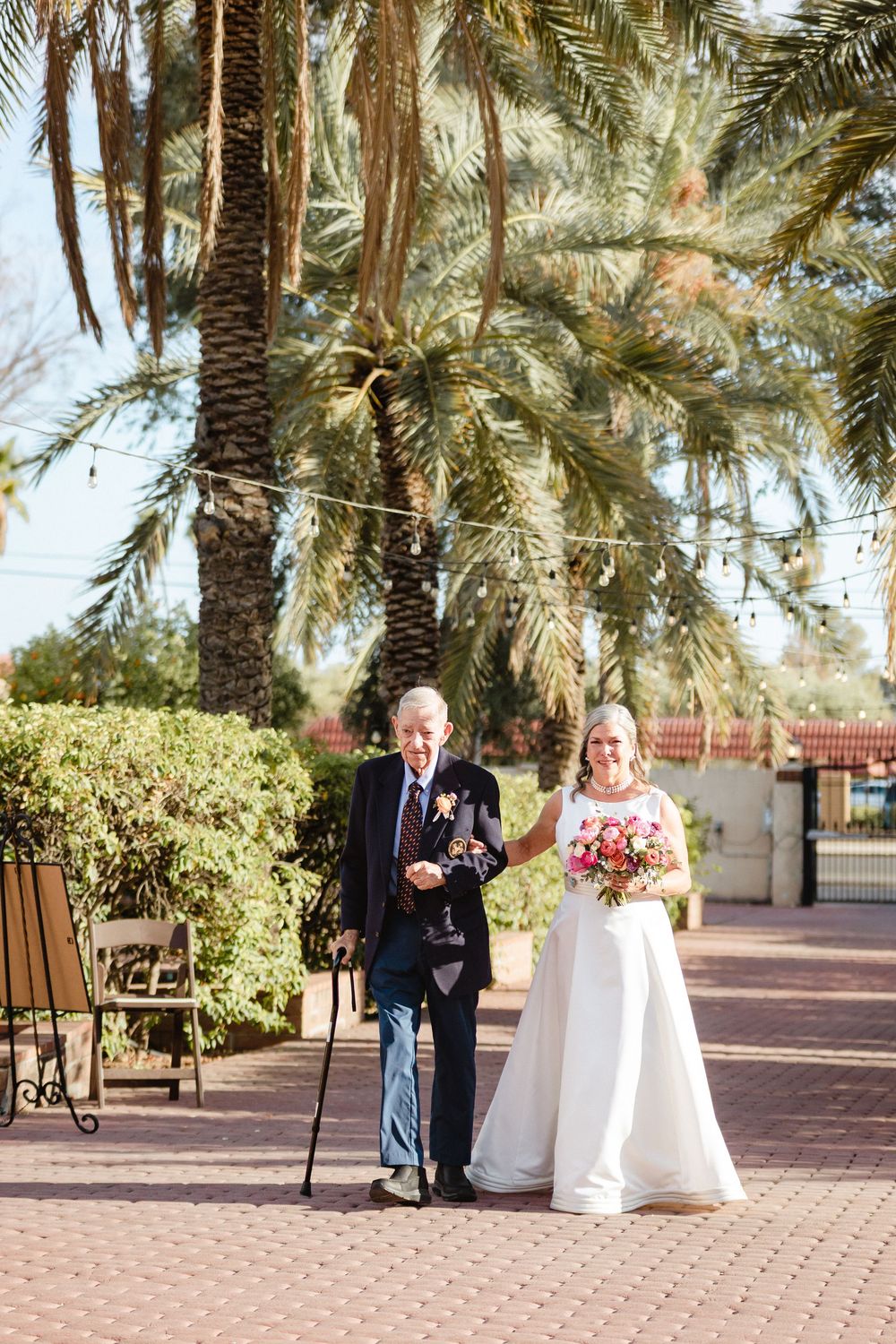 Wedding processional on palm tree-lined brick path with bridal bouquet under bright sunlight in outdoor venue.