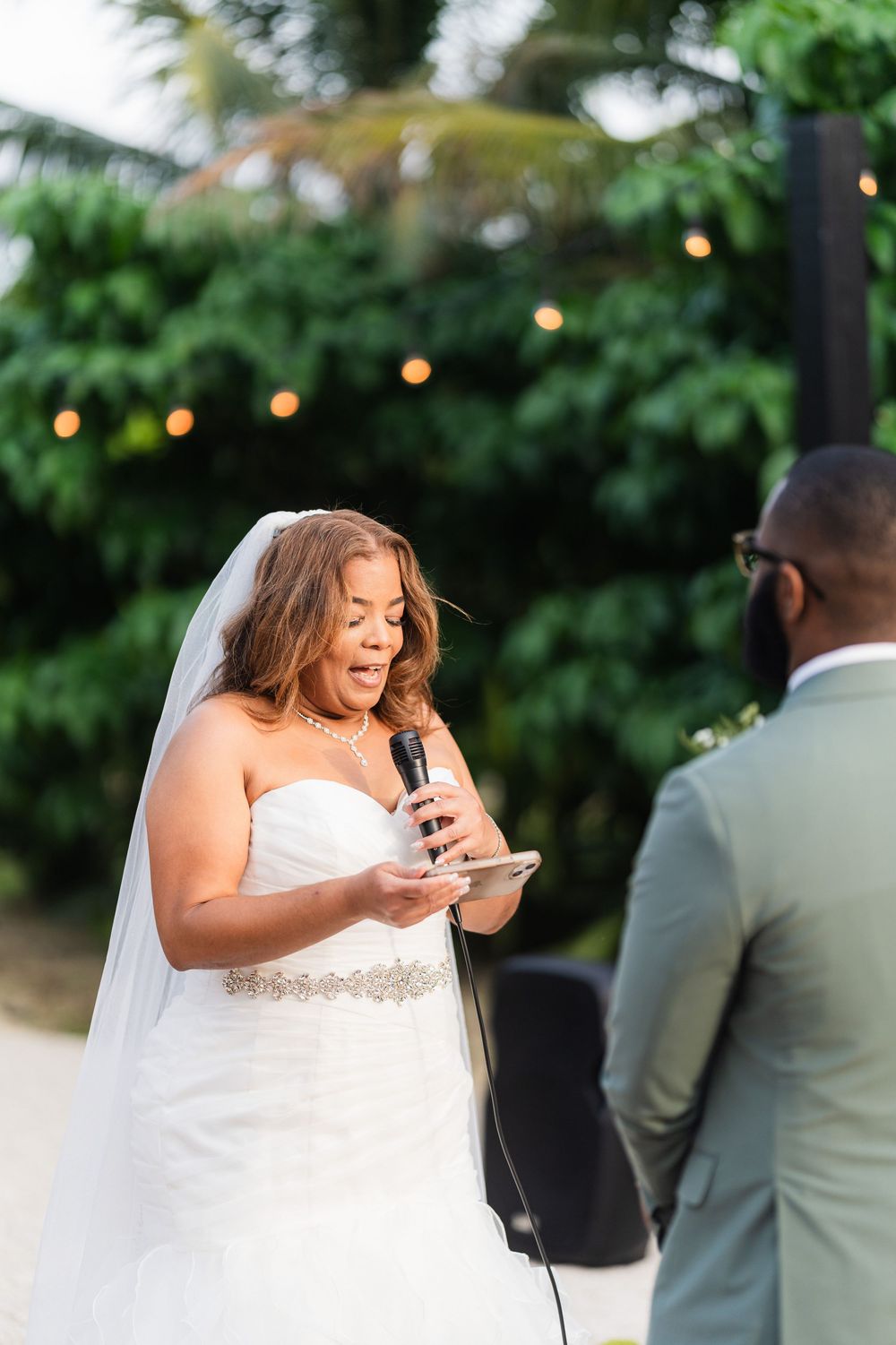 Bride in white dress reads vows with emotional expression during outdoor ceremony.