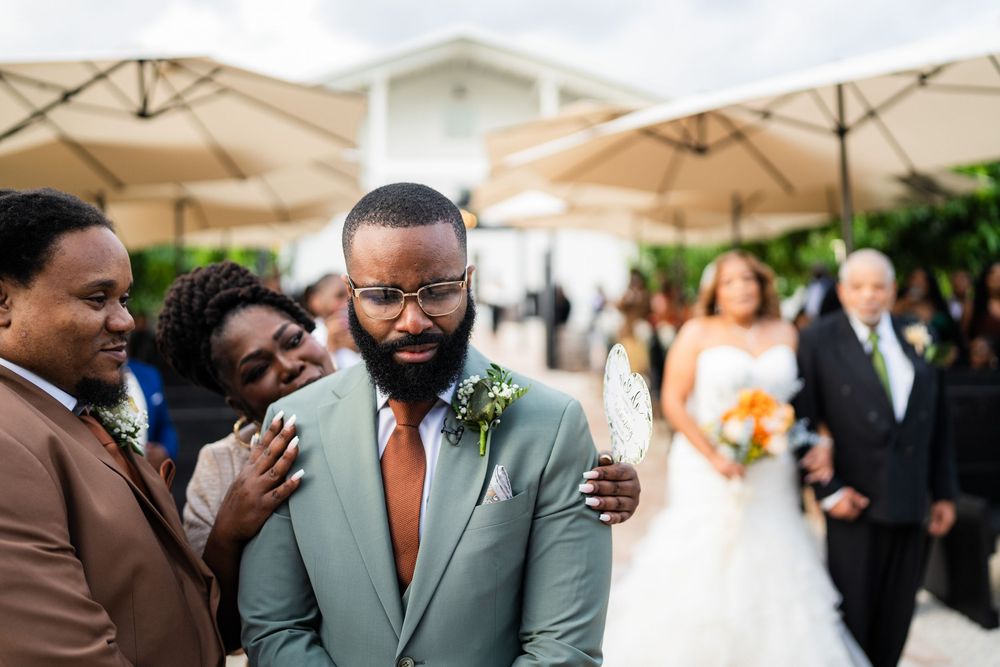 A group of formally dressed wedding guests comfort and embrace an emotional groom during an outdoor celebration.