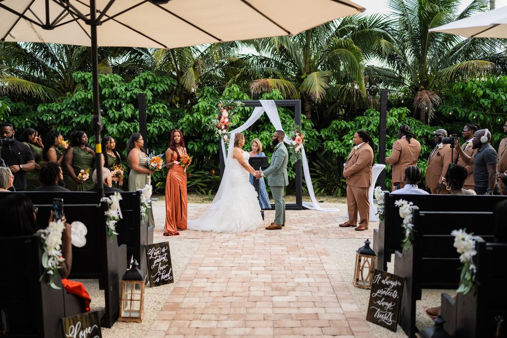 A wedding ceremony takes place under a decorated arch on a brick patio surrounded by tropical palm trees and guests.