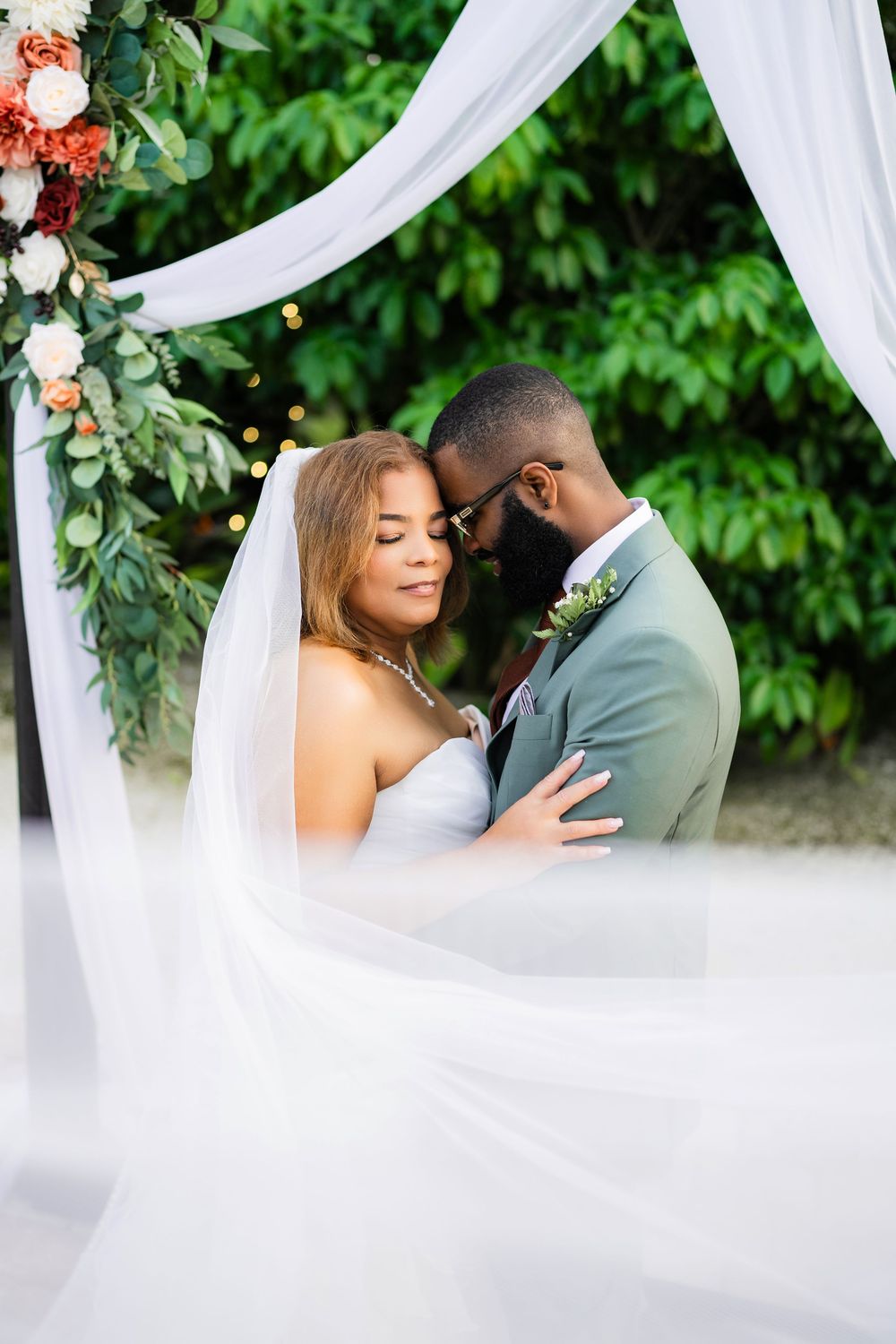 A romantic wedding moment captured under a white draped arch decorated with coral flowers and greenery.