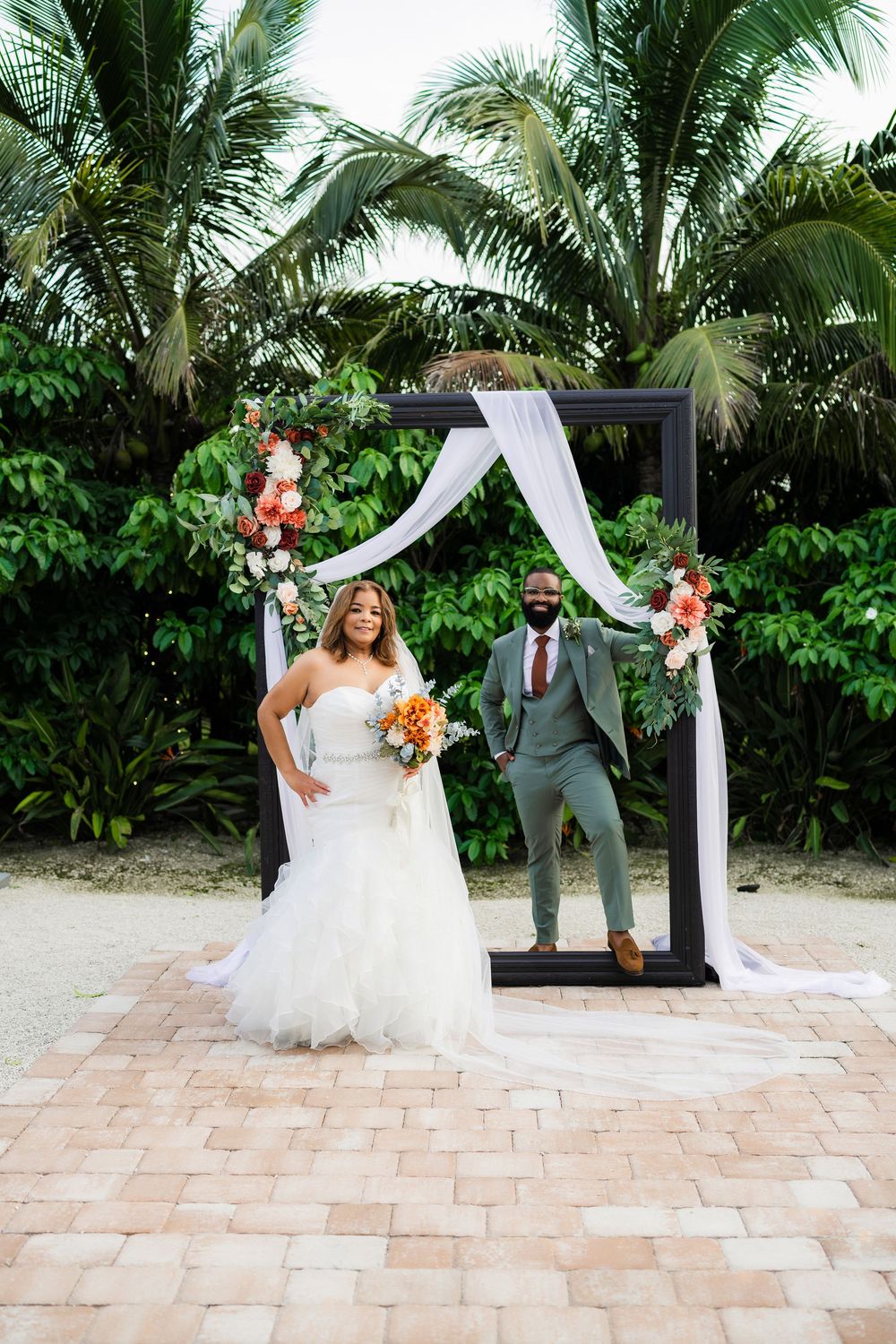 Wedding couple poses under a modern black arch decorated with white fabric and coral flowers against tropical palm trees.
