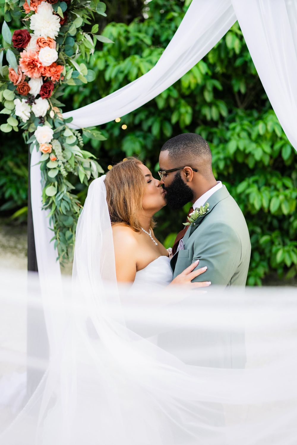 Intimate wedding portrait of newlyweds embracing under a floral arch with flowing white fabric.