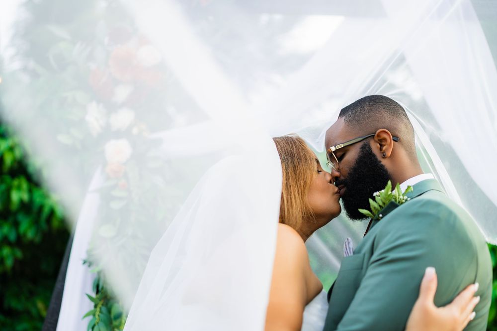 A couple shares a romantic kiss under a flowing white veil during their outdoor wedding ceremony.