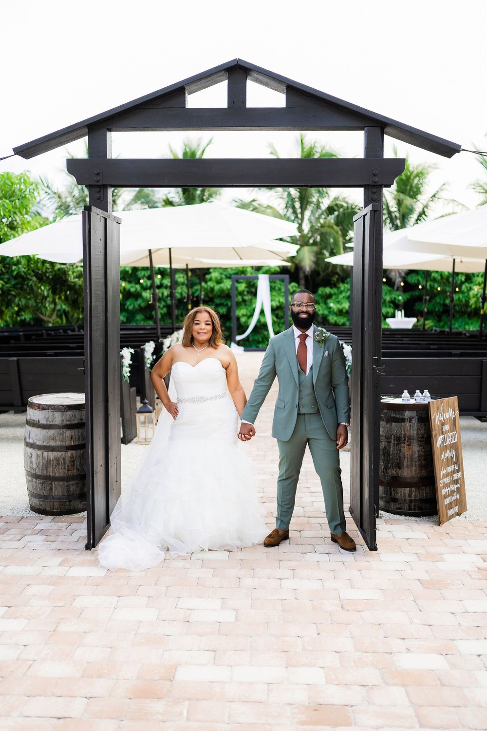 A joyful wedding couple stands beneath a black wooden pergola with rustic barrels and greenery.