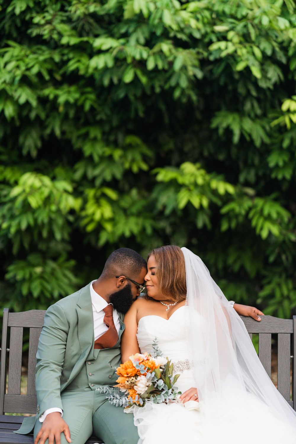 Newlyweds in sage green and white share a tender moment on a garden bench with their bridal bouquet.