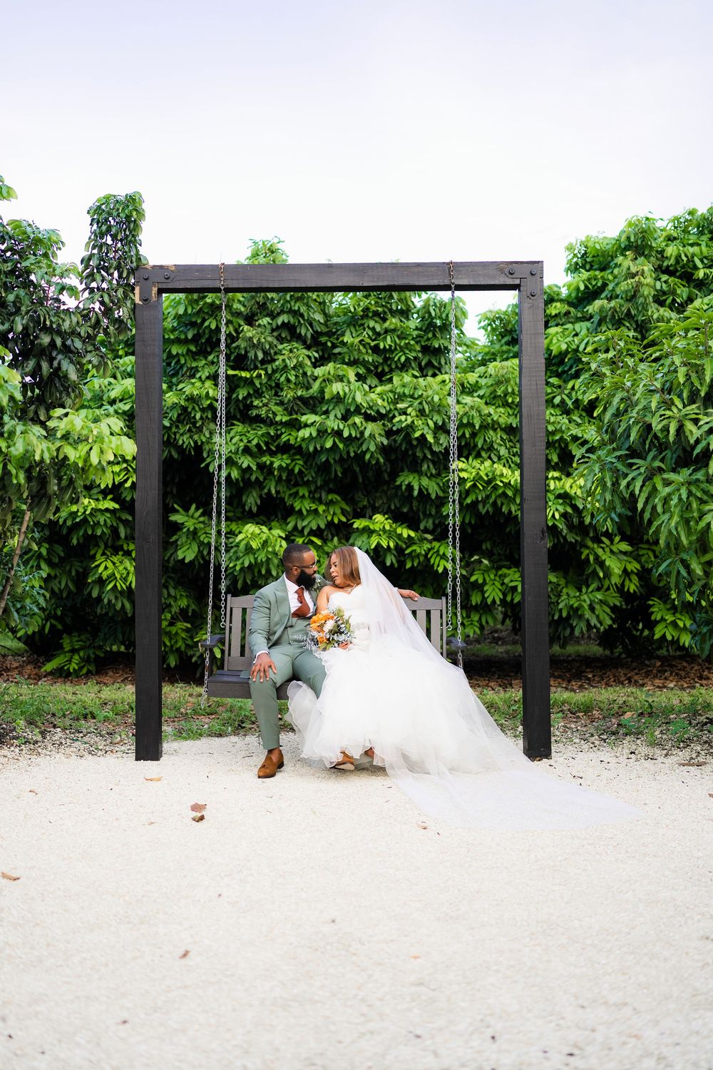 Wedding couple sits together on a modern black metal swing against lush green garden backdrop.