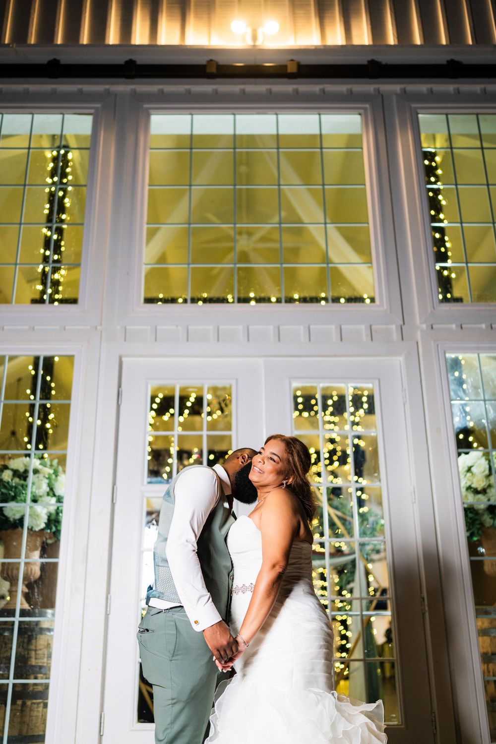 Newlywed couple embraces in front of elegant white French doors decorated with glowing string lights at wedding venue.