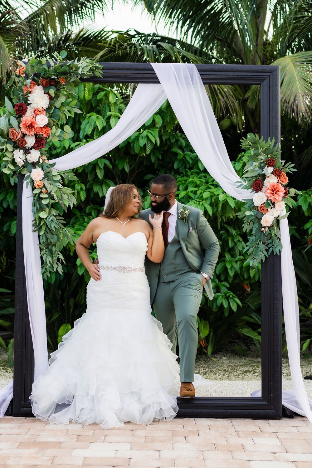 A wedding couple poses under an elegant black arch decorated with white fabric and coral floral arrangements in a tropical garden.