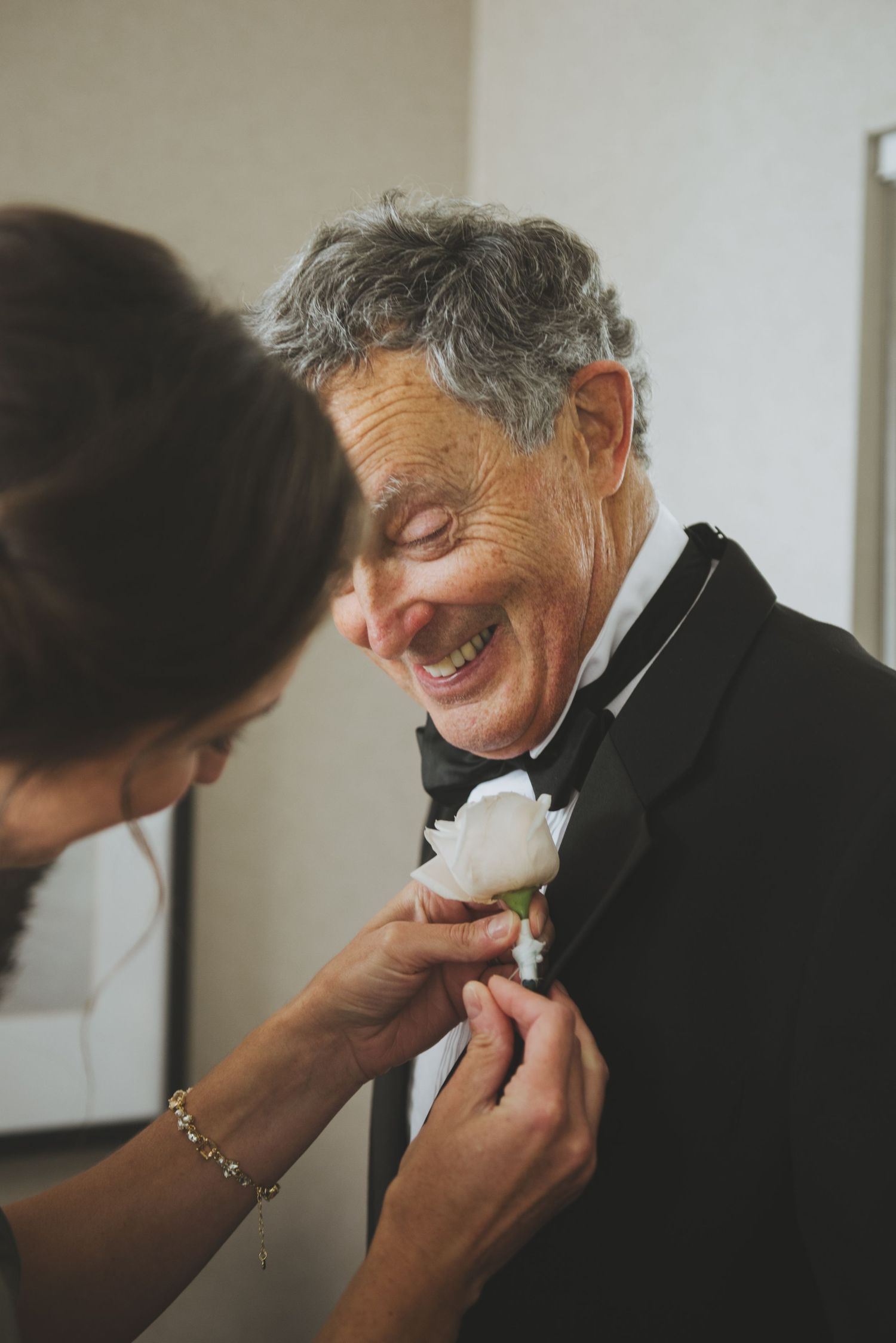Someone helps attach a white rose boutonniere to a man's formal black tuxedo lapel while he smiles joyfully.