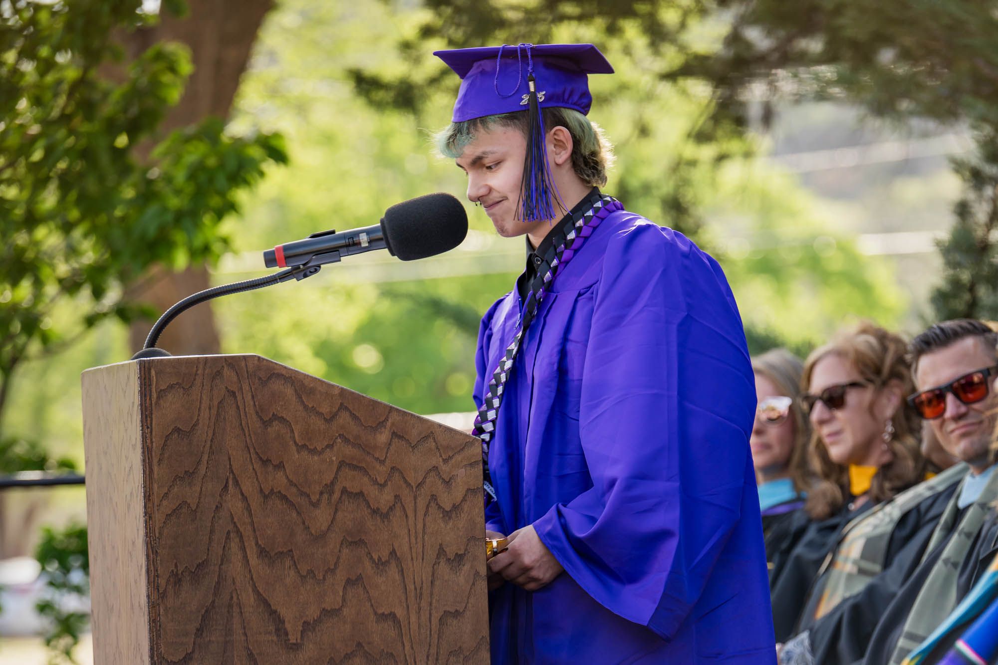 Graduate in royal blue cap and gown gives speech at outdoor ceremony podium while audience watches attentively.