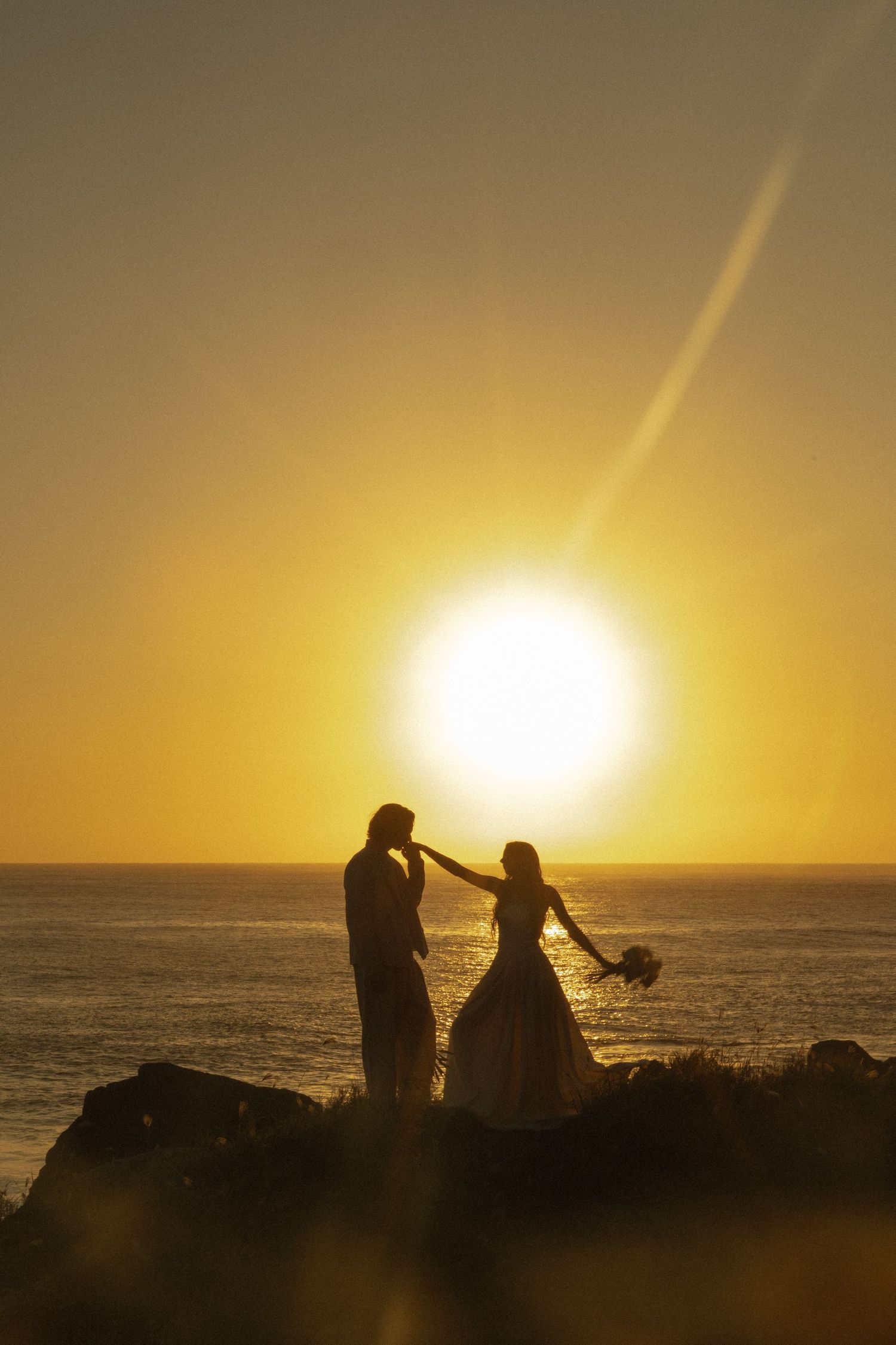 Two people holding hands walk along a coastal cliff path at sunset with ocean views and dramatic clouds in the background.