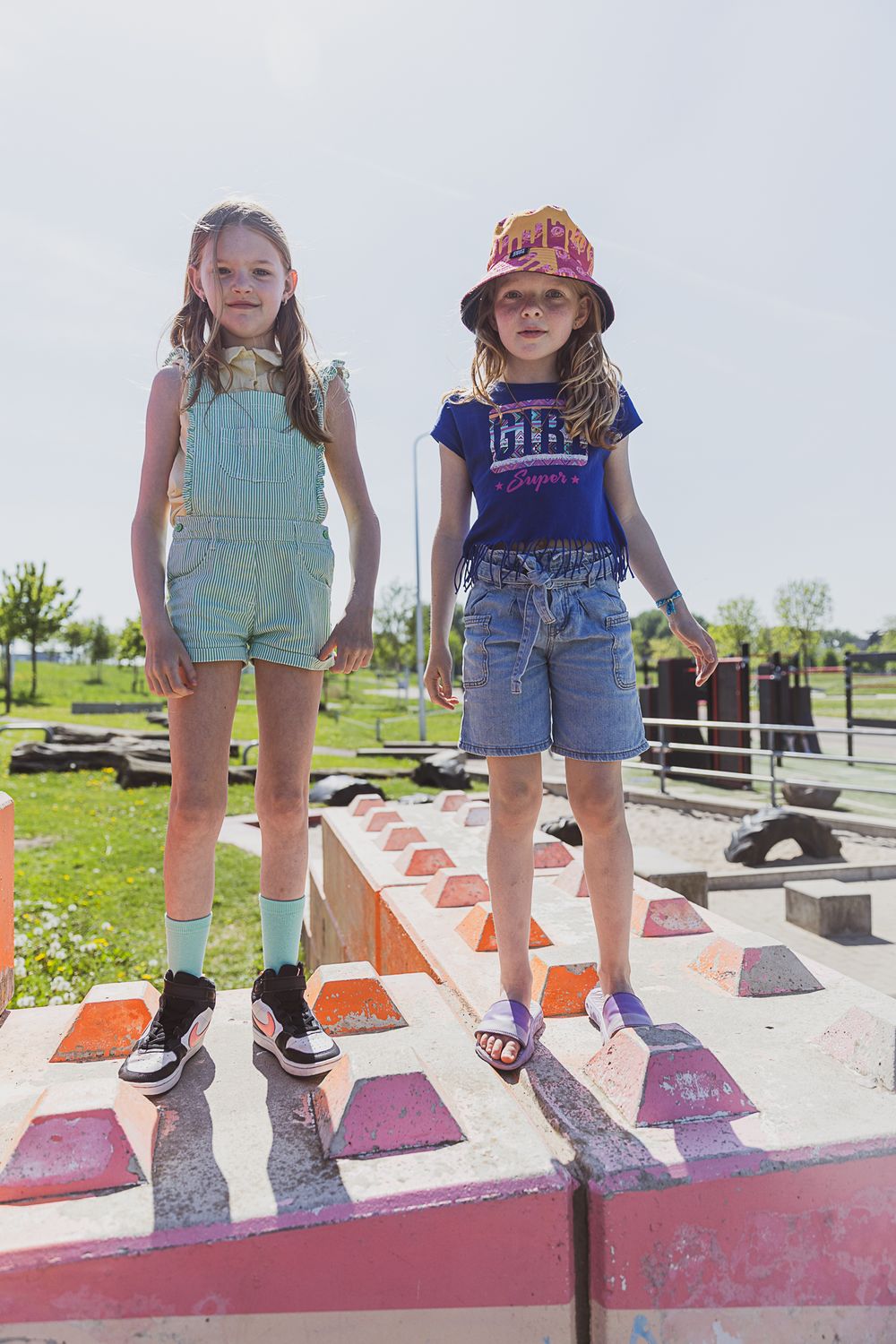 Two children stand on a colorful painted playground structure on a sunny day wearing summer casual outfits.