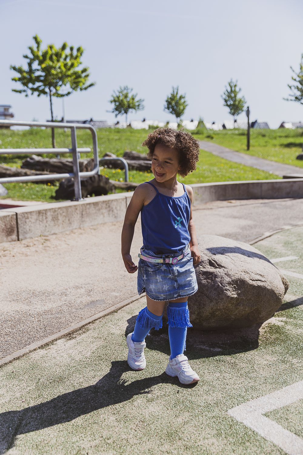 Child in blue tank top and denim shorts enjoys a sunny day in a park with trees and rocks.