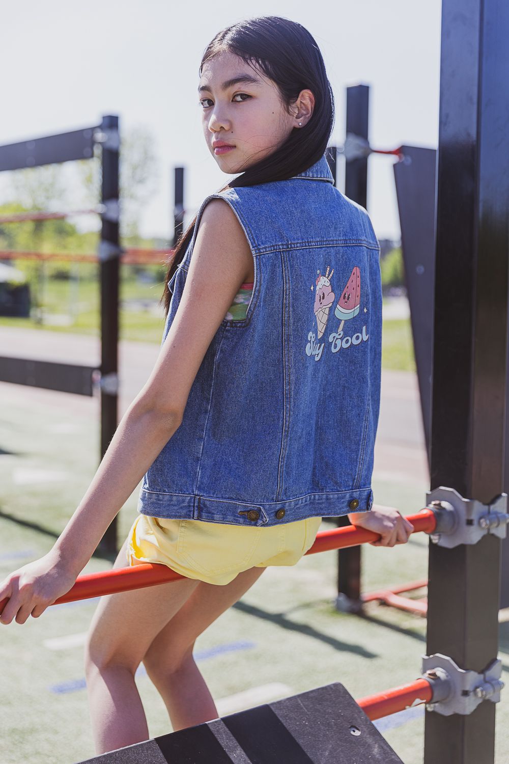 Young person wearing denim vest with butterfly design poses on outdoor playground equipment in sunny setting.