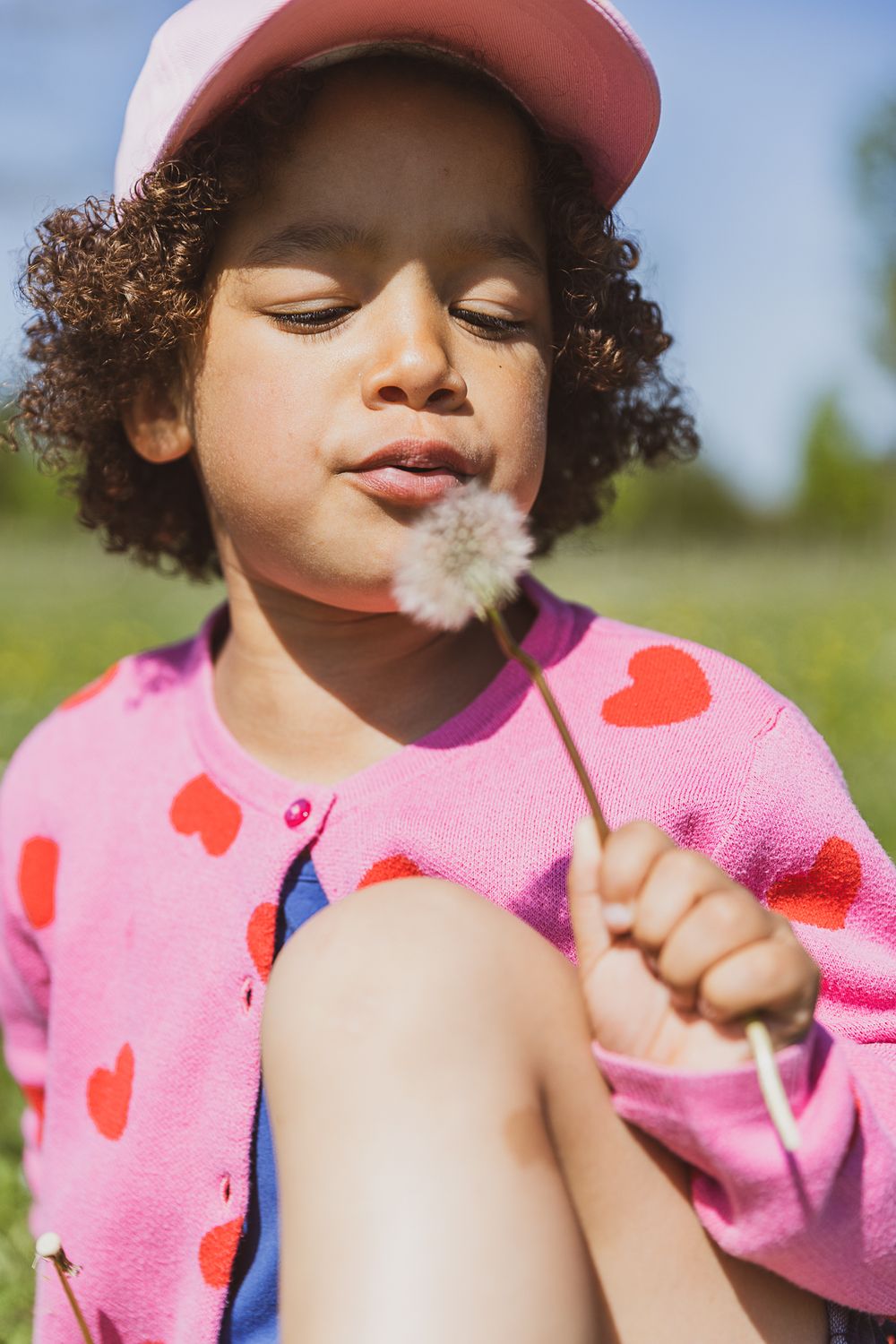 Child wearing pink heart-patterned sweater and cap gently blows on a dandelion in the sunlight.