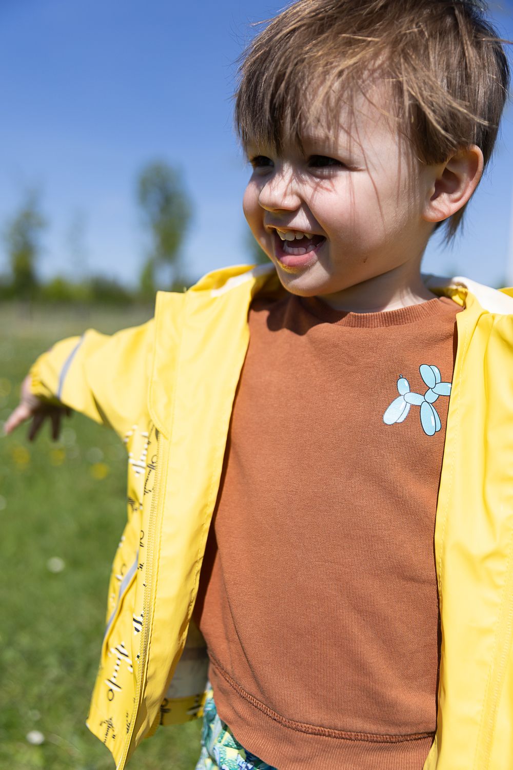 Young child smiling outdoors wearing a brown shirt and yellow jacket on a sunny spring day in a grassy field.