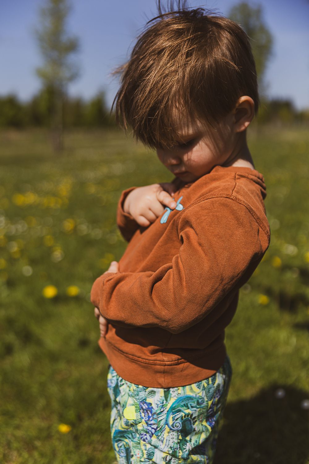 Small child wearing an orange sweater and floral pants stands in a sunny meadow with yellow wildflowers.