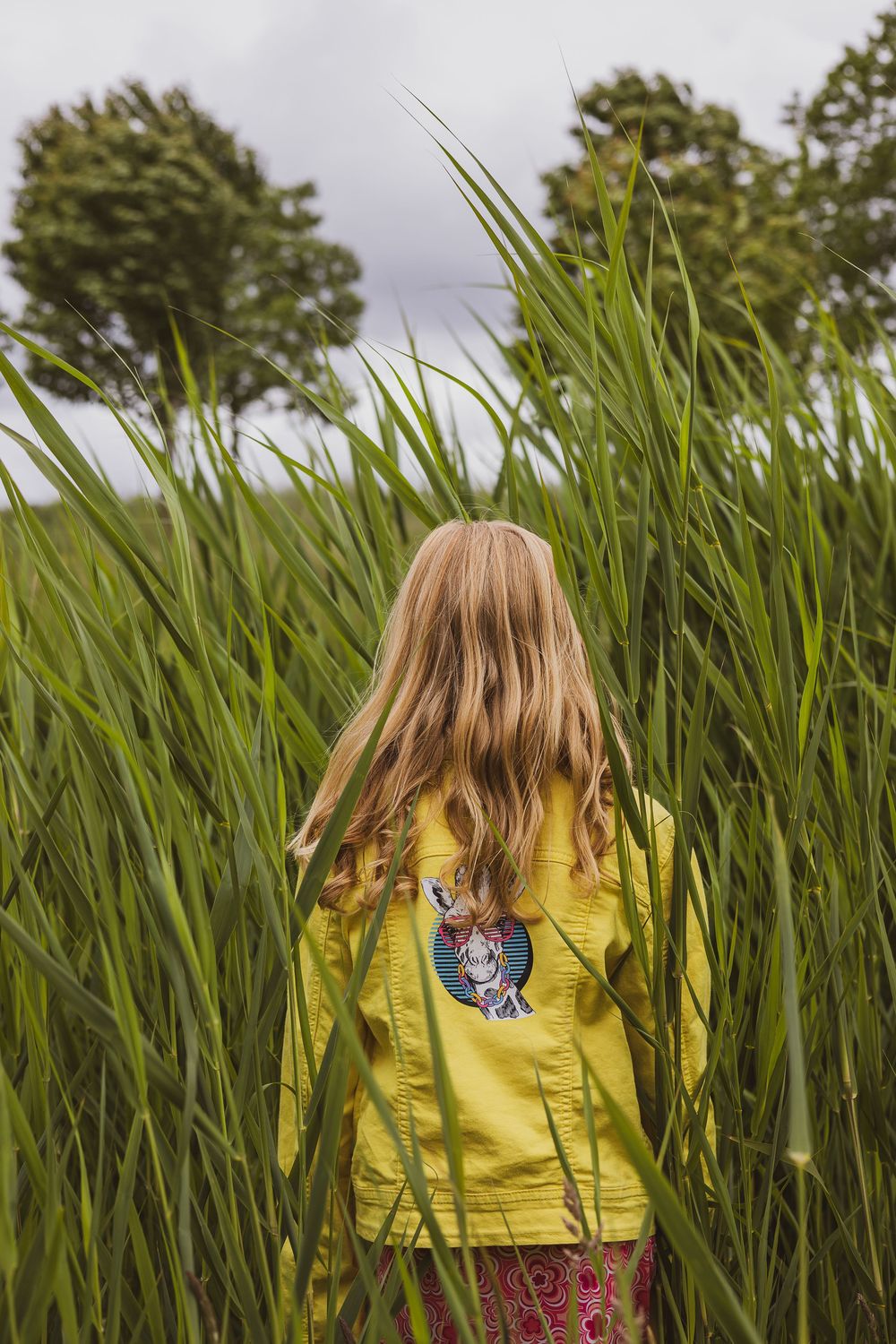 A young person wearing a bright yellow jacket stands in tall green grass on an overcast day.