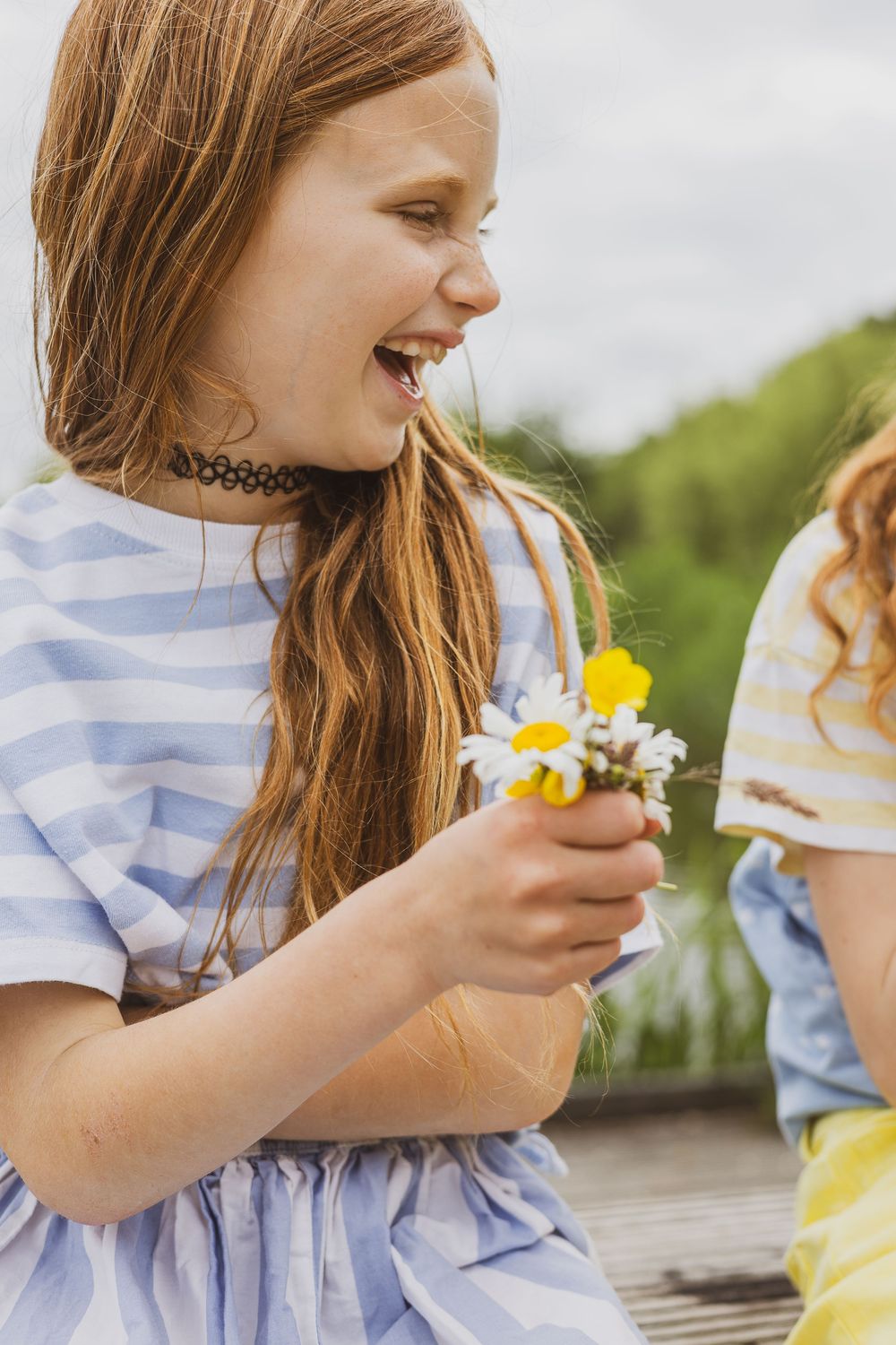 Young person in striped blue dress laughing joyfully while holding fresh picked daisies and wildflowers outdoors.