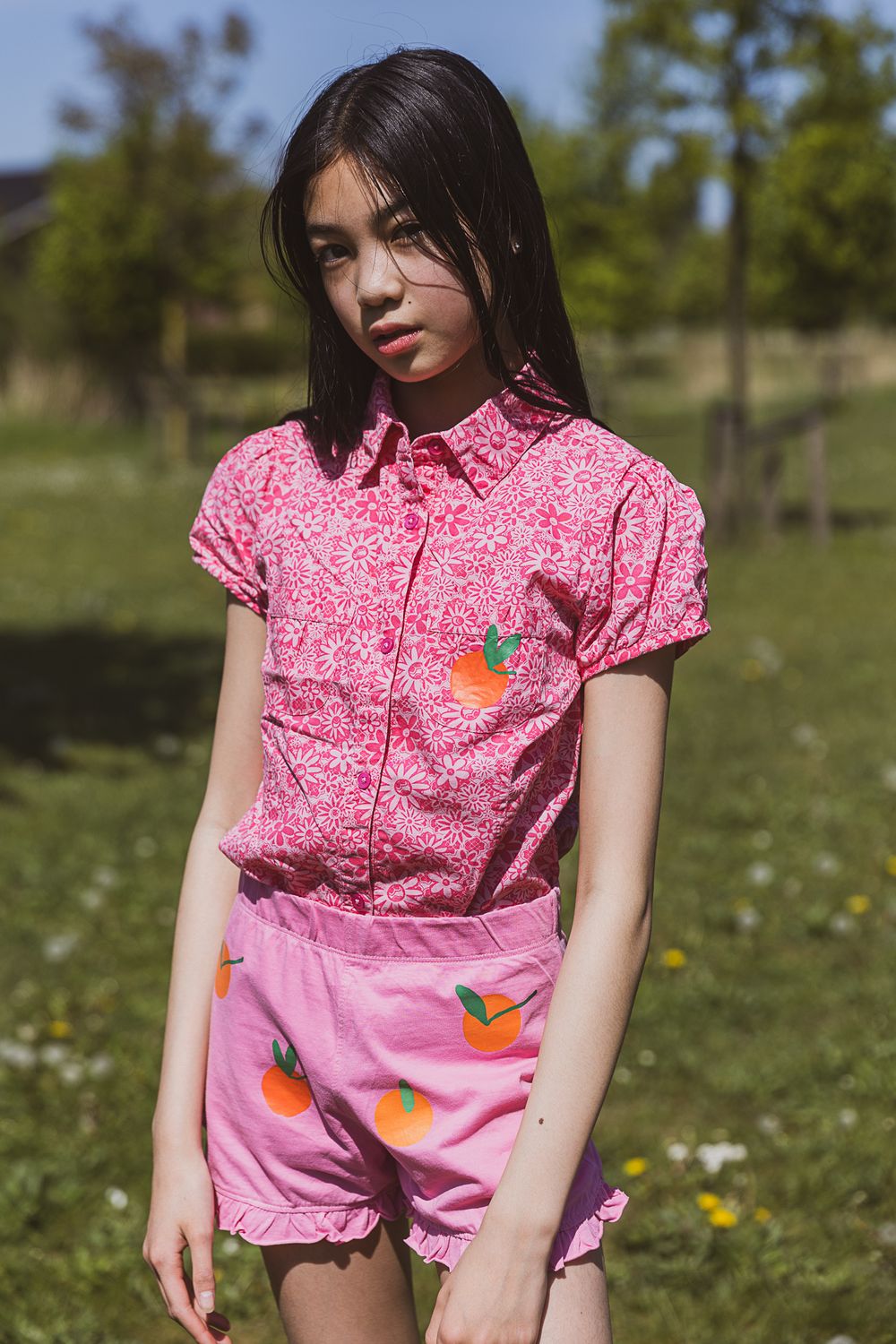 Person wearing pink floral button-up shirt and matching pink shorts stands in sunny meadow with dandelions and trees.