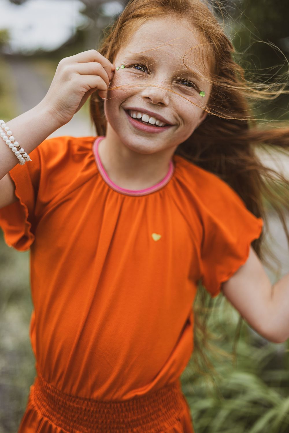 Child wearing bright orange clothing with flowing hair smiles joyfully while playing outdoors.