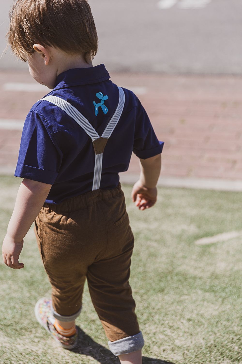Back view of a toddler in navy polo shirt and brown pants with suspenders walking on grass.