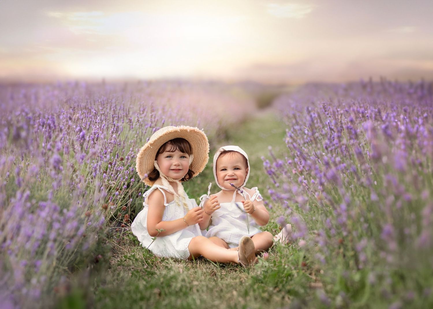 Two children sit together in a dreamy lavender field during sunset wearing white dresses and straw hats.
